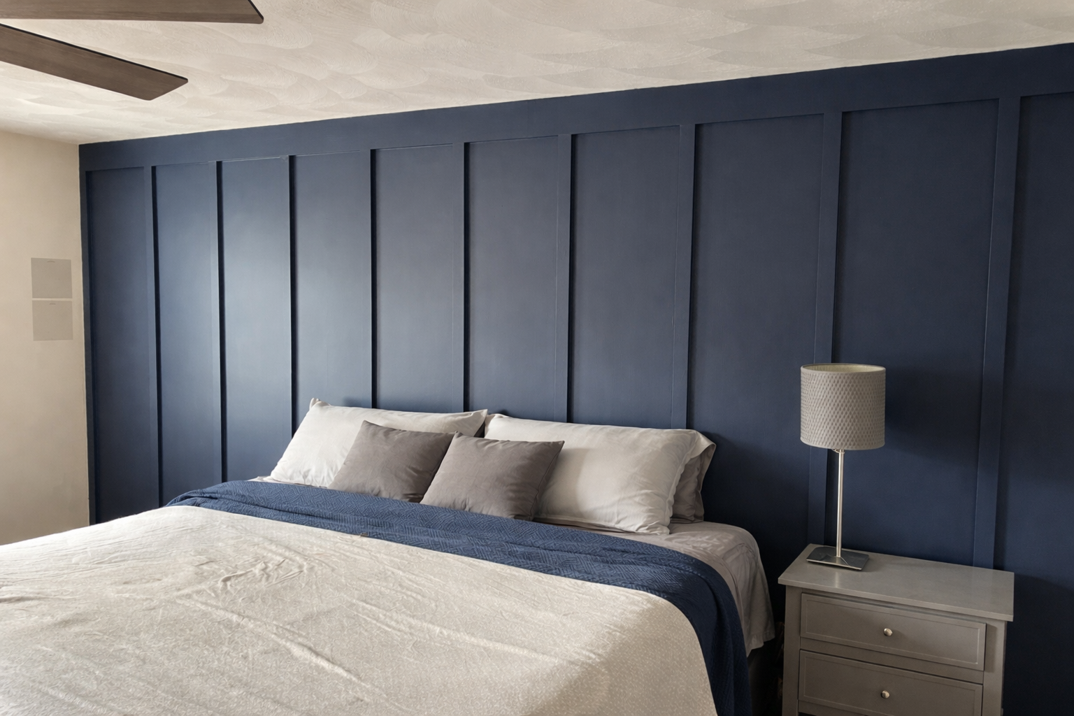 A bedroom with a large bed covered with white bedding and gray pillows, a dark blue paneled accent wall behind the bed, and a white bedside table with a silver lamp to the right.