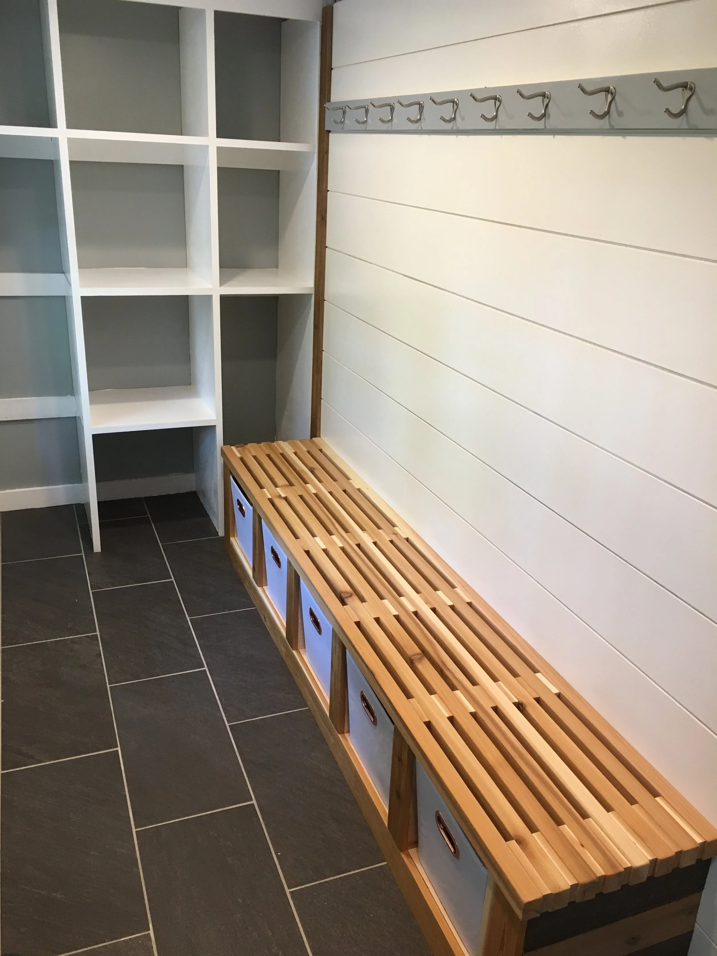Entryway or mudroom with black tile flooring, white shelving on the left, and a wooden bench with blue storage bins underneath on the right wall with hooks above.