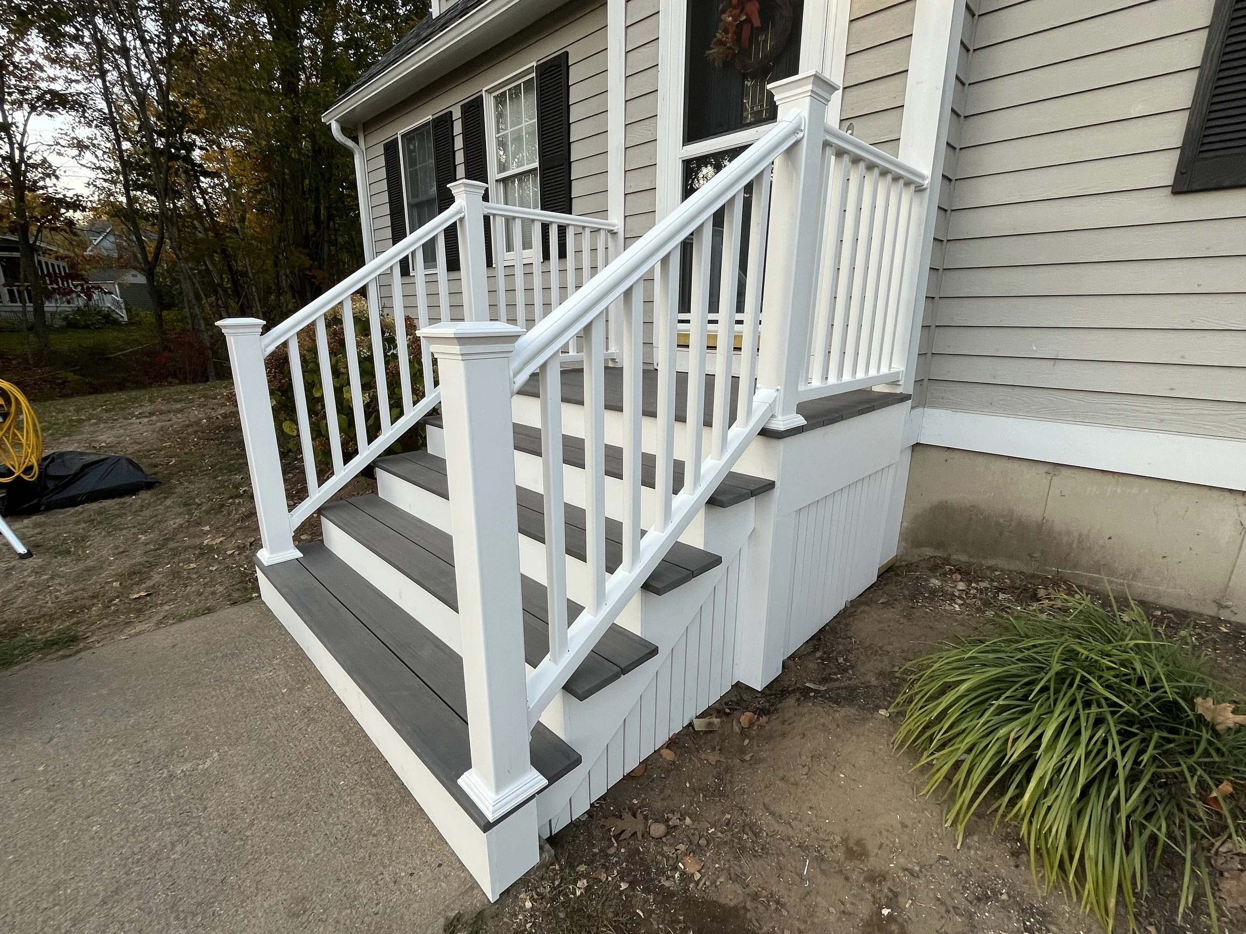 Newly constructed white wooden staircase with gray steps leading up to a house porch, with black shutters on windows, situated next to a gardening bed with green foliage.