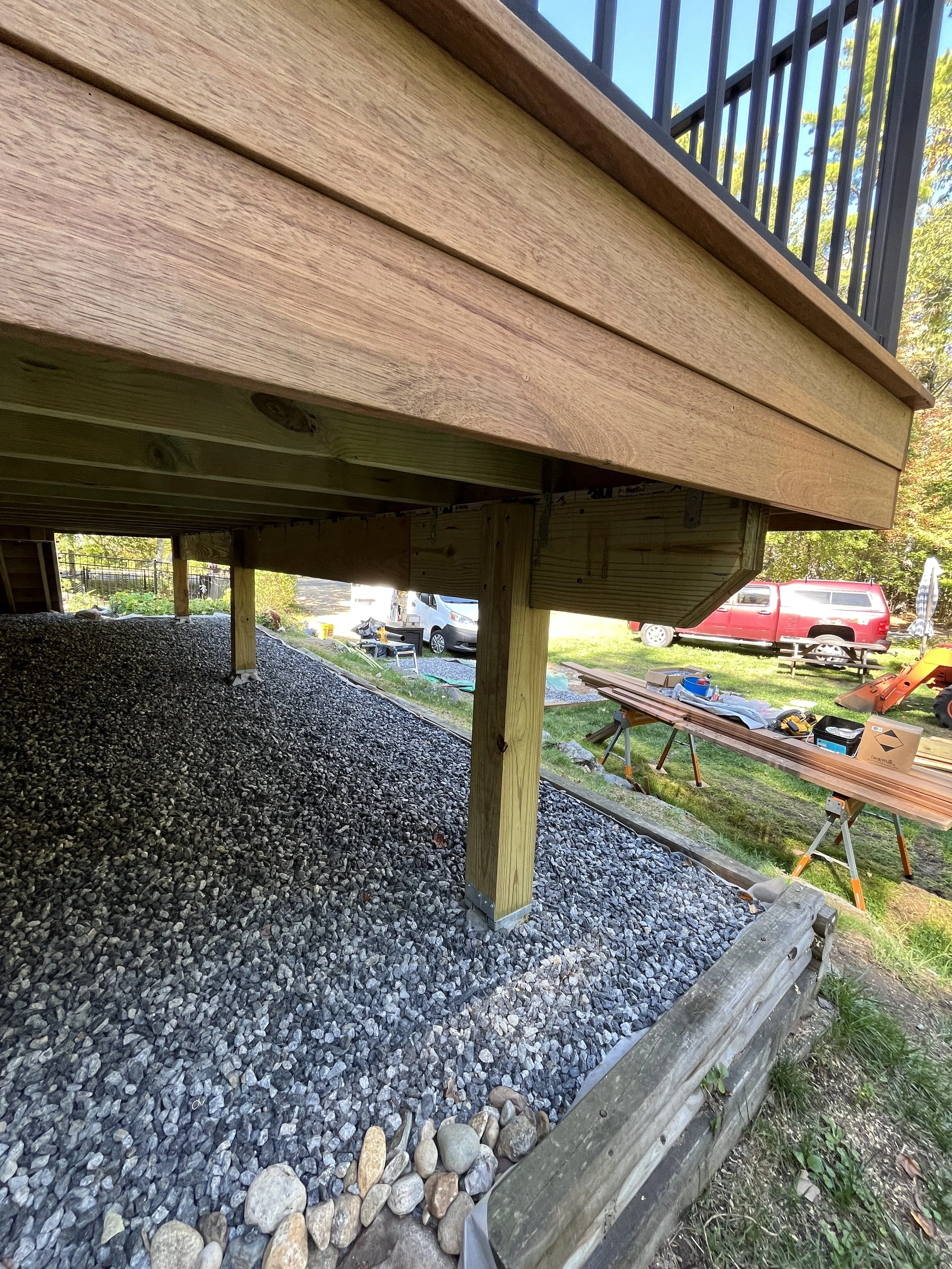 Underside view of a wooden deck with support posts and gravel foundation, with construction tools and materials visible in the background.