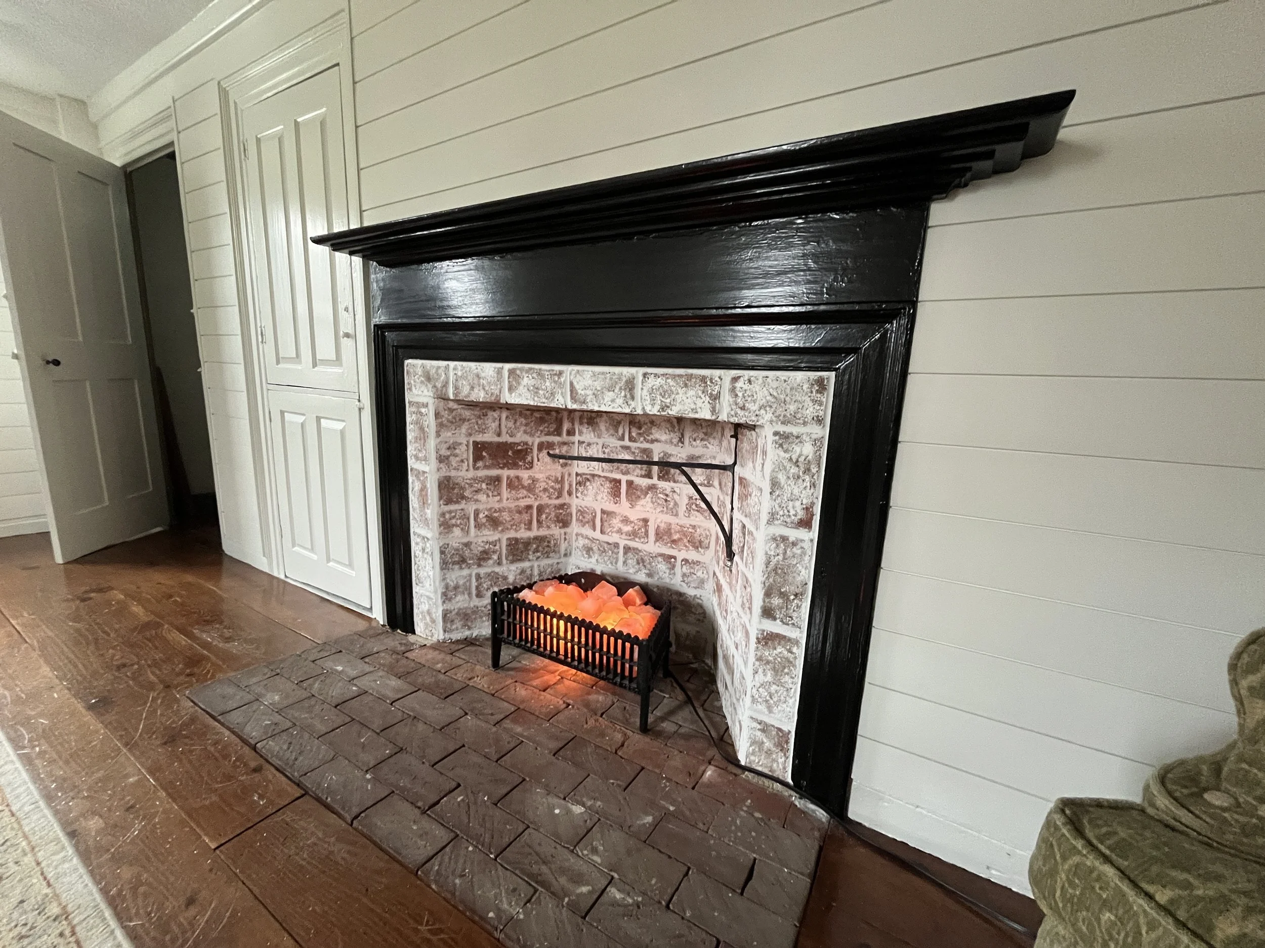Living room fireplace with black mantel, brick interior, and salt lamp in a wire basket, hardwood floor, and partially visible armchair.