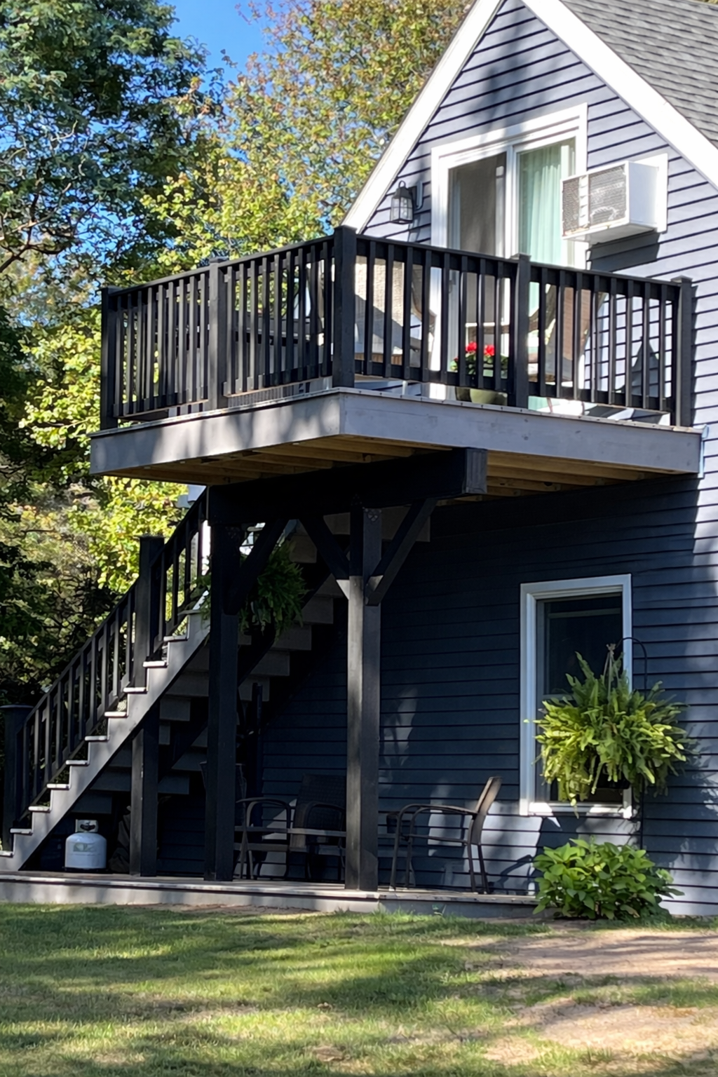 Two-story house with blue siding and white trim, featuring a small upper balcony with black railing and a lower patio area with chairs, surrounded by greenery and trees.