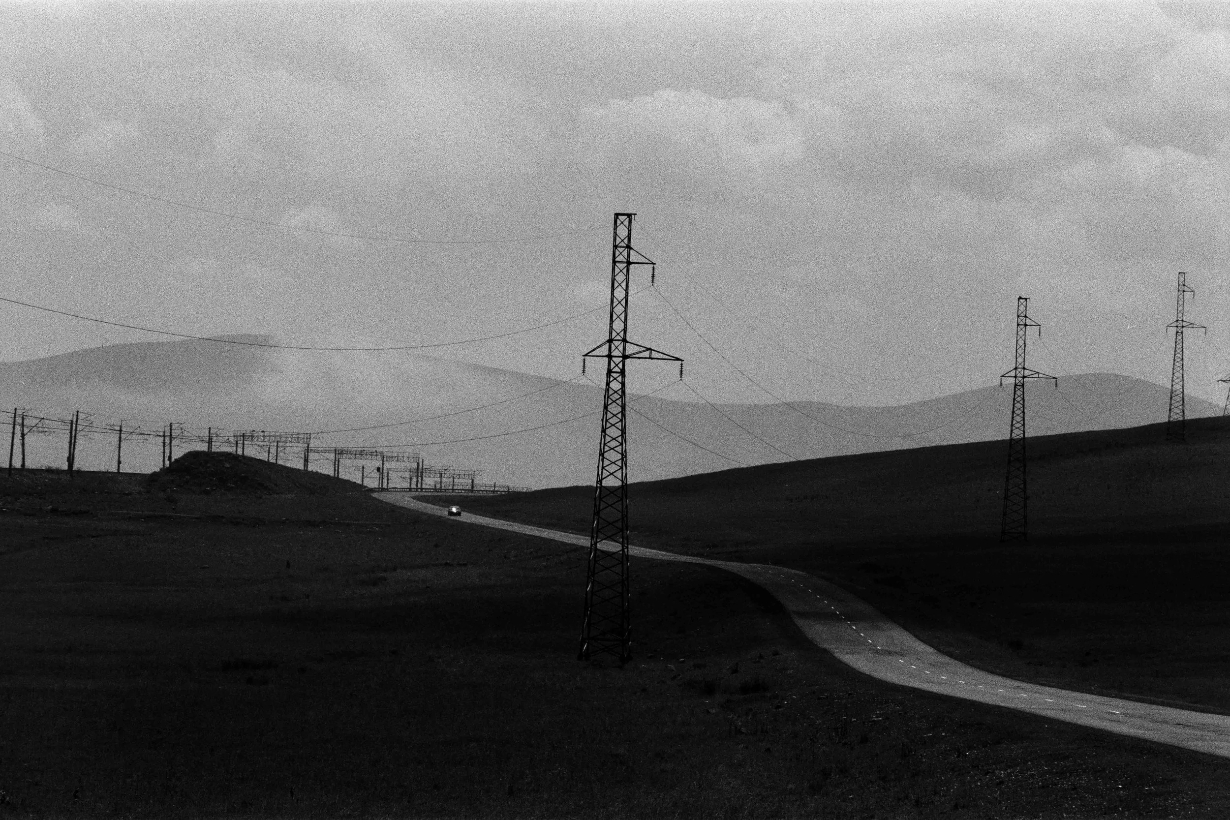 Paysage rural avec une route sinueuse, plusieurs pylônes électriques, et des collines en arrière-plan, en noir et blanc.
