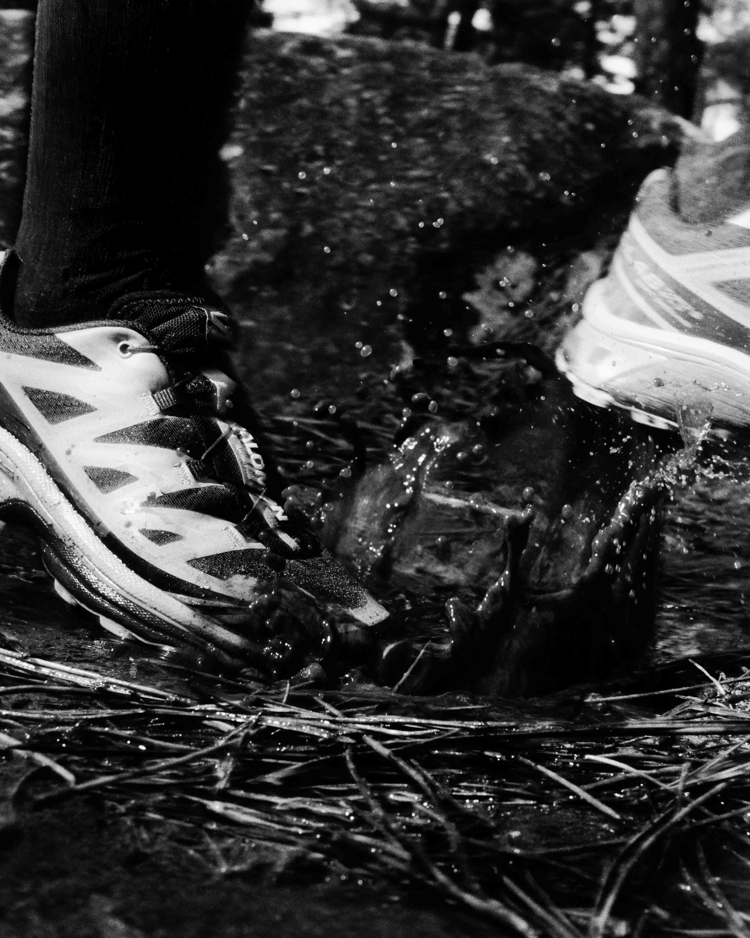 Chaussures de course en train de fouler un terrain mouillé avec de l'eau et des branches, photo en noir et blanc.