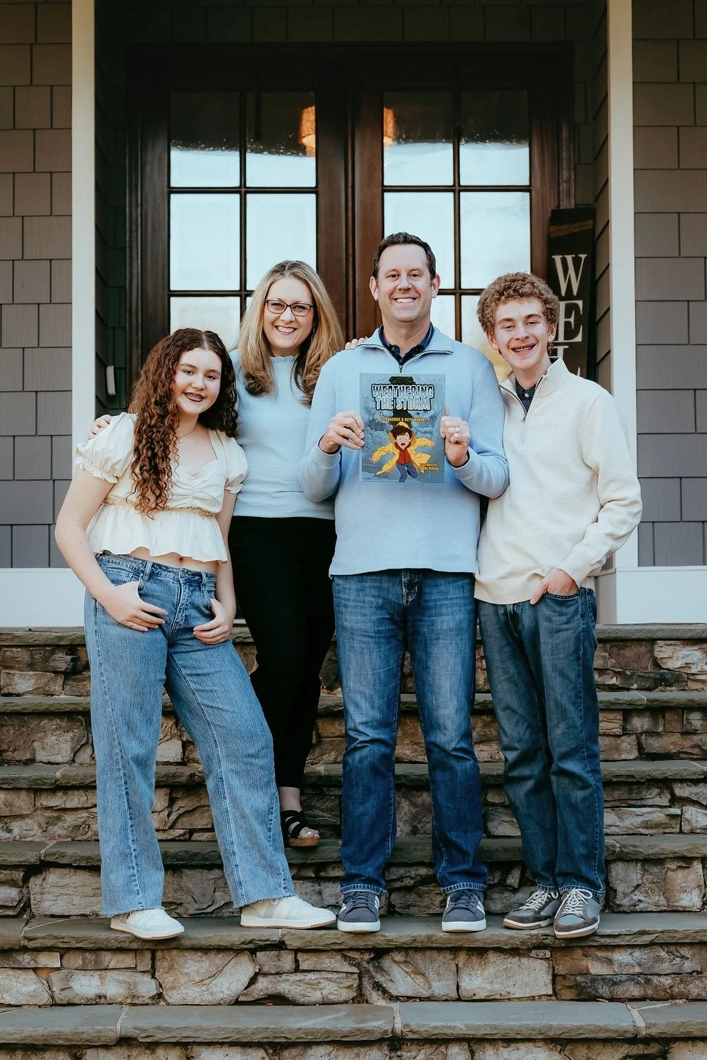 A family of four stands on the steps of a house, posing for a photo. The father holds a book titled 'Weathering The Storm'.