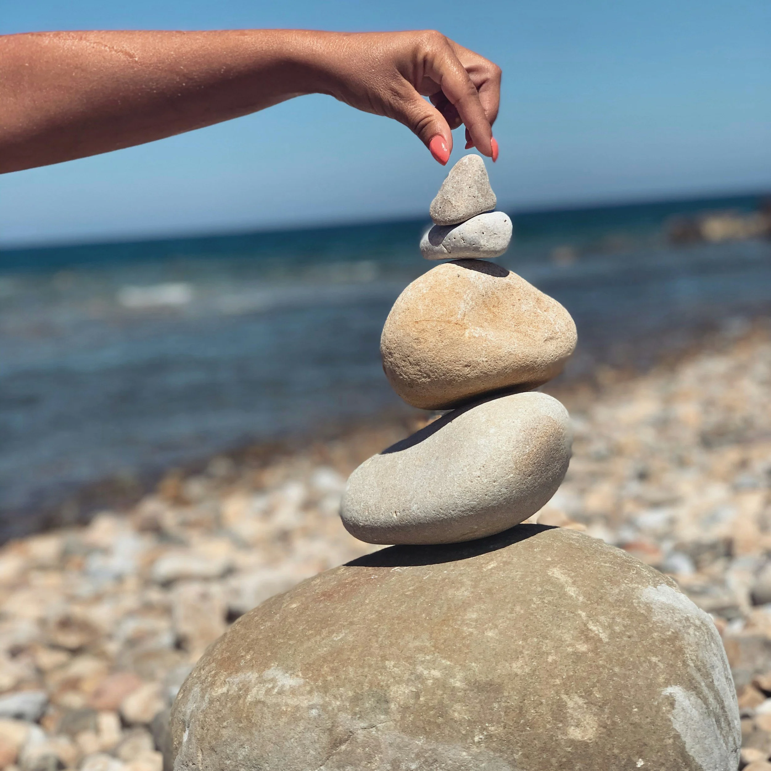 A person's hand balancing five stacked stones of varying sizes on a large base stone on a beach with the ocean in the background.