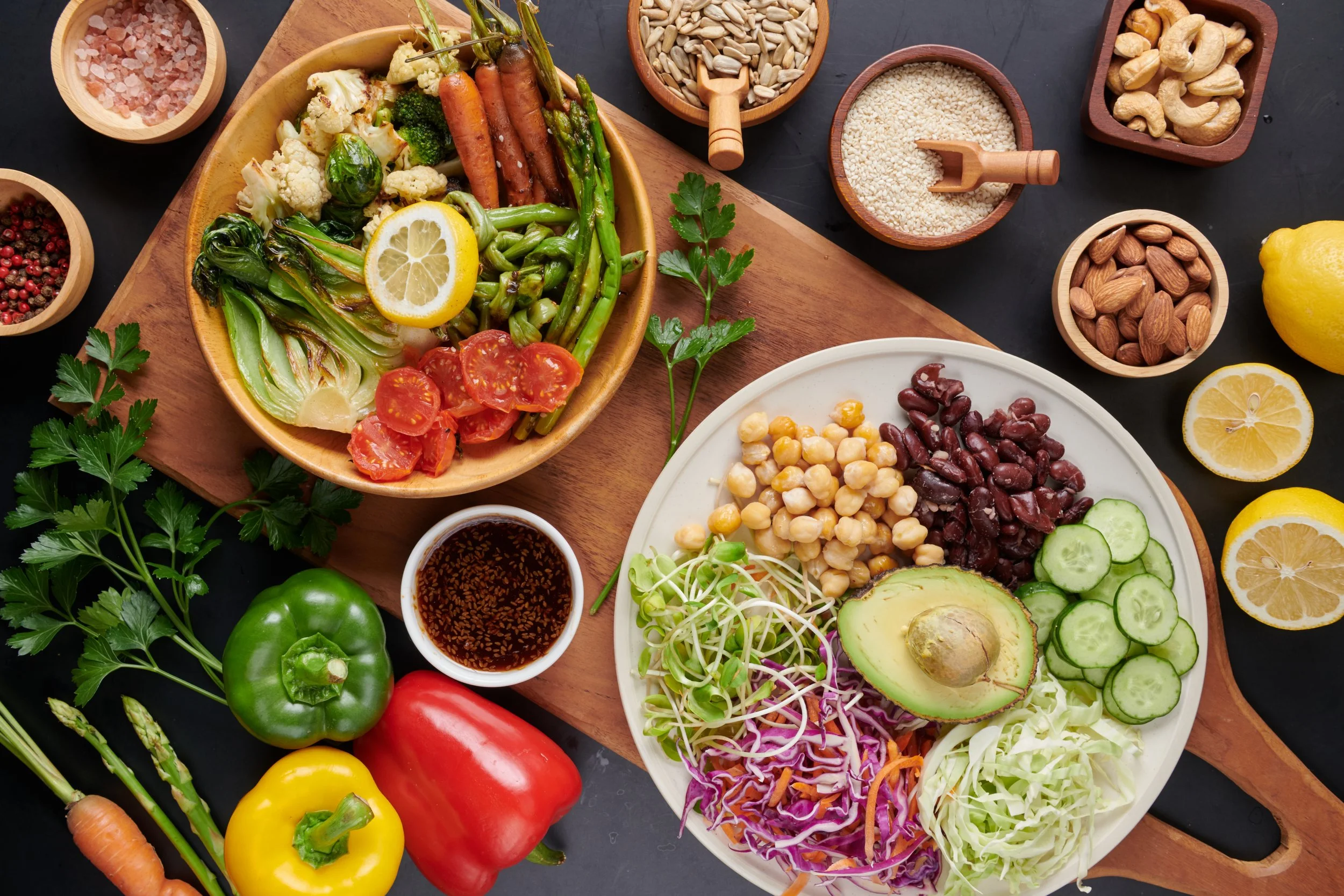 A variety of fresh vegetables, beans, grains, nuts, lemon slices, and a small bowl of dressing or sauce arranged on a dark surface for a healthy salad meal.