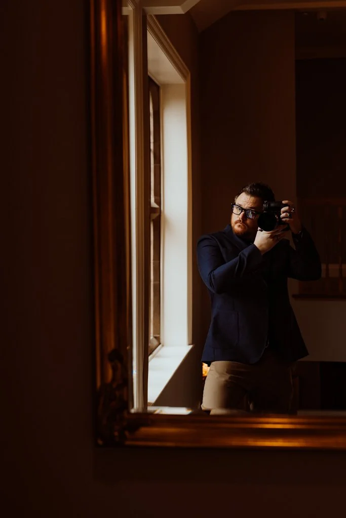 Man taking a selfie in a mirror near a window, wearing glasses, a dark blazer, and khaki pants.