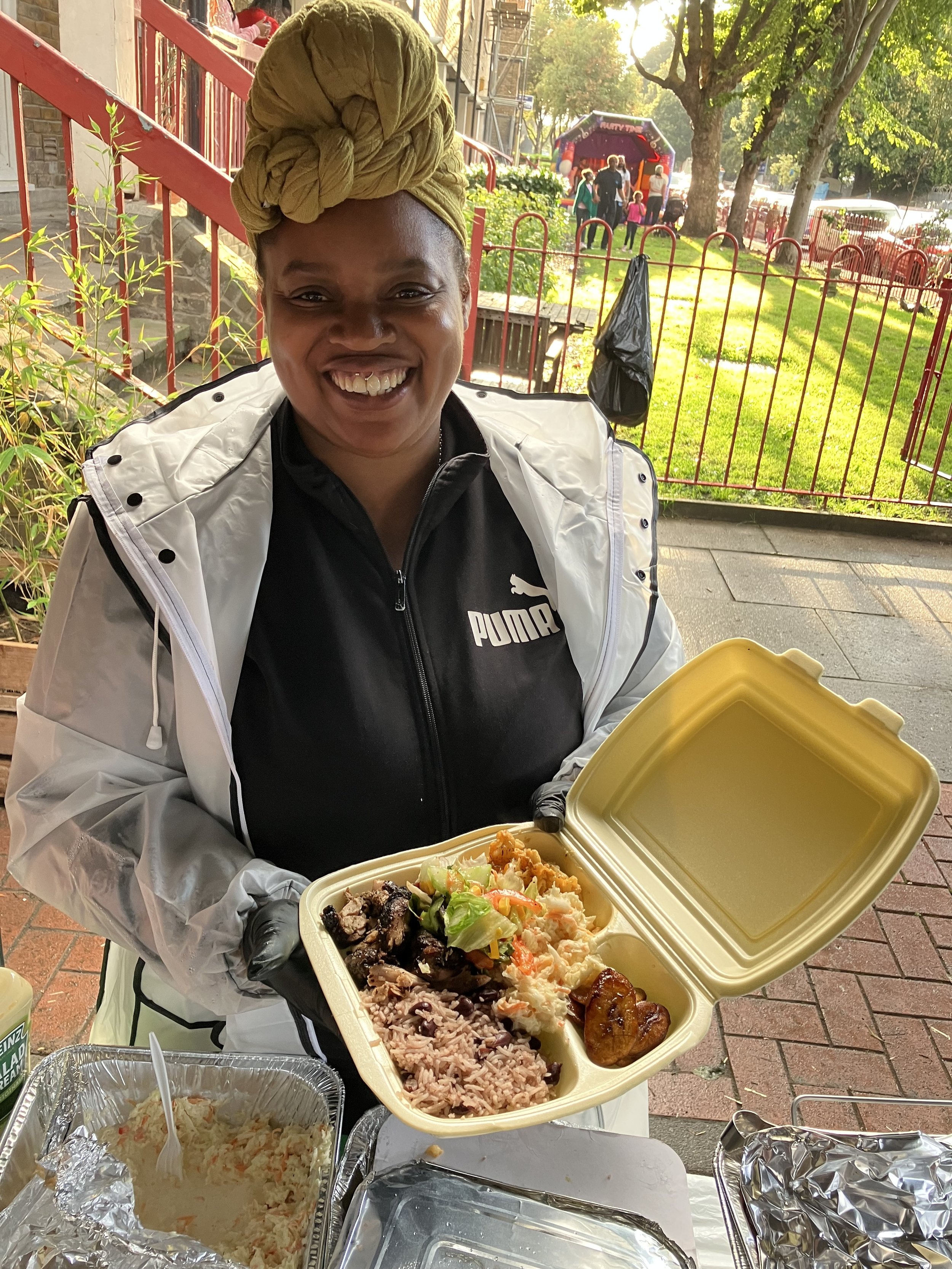A woman with a yellow head wrap, black gloves, and a black Puma jacket smiling and holding an open takeout food container with rice, potato salad, greens, meat, and a fried item, in an outdoor setting.