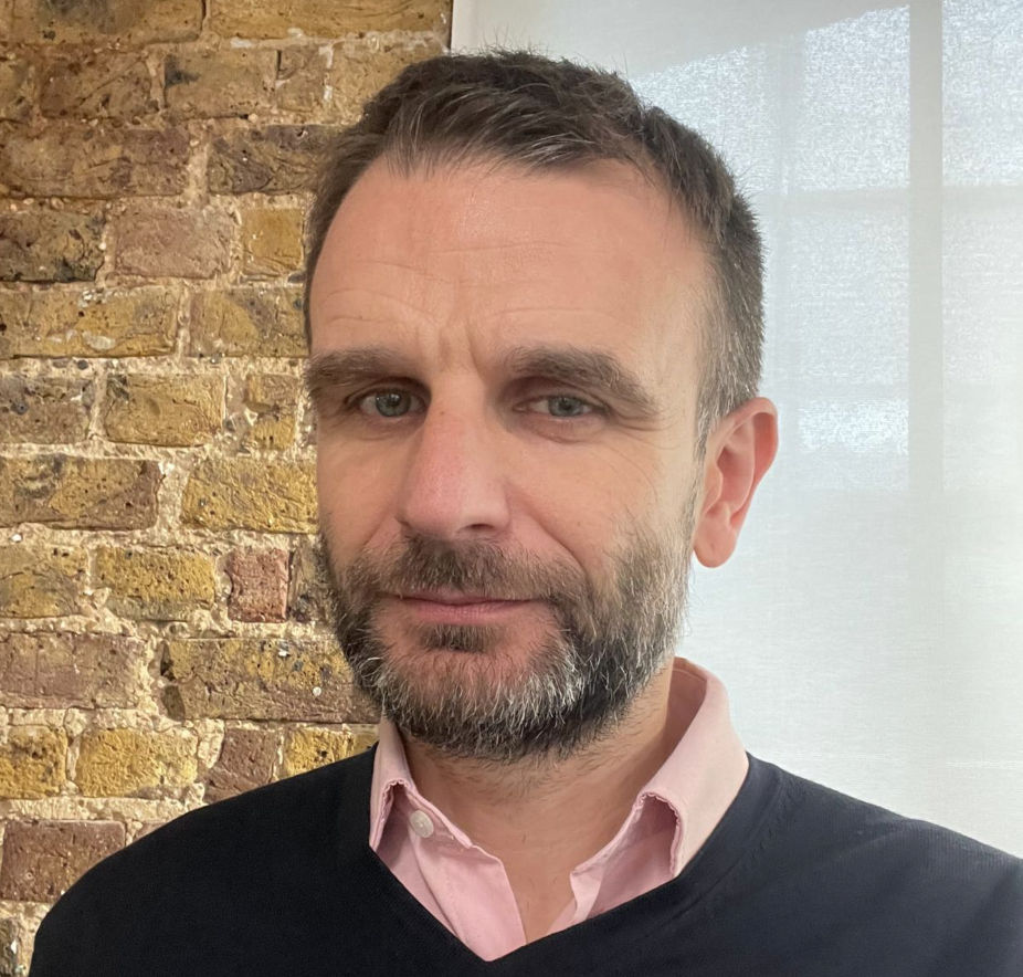 A middle-aged man with short brown hair and a beard, wearing a light pink collared shirt under a black sweater, standing indoors in front of a brick wall and a window with white curtains.