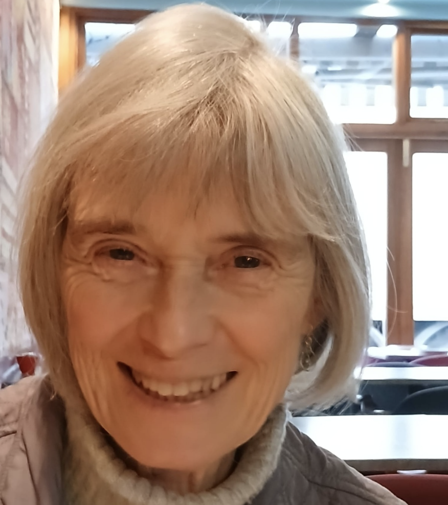 Close-up of a smiling older woman with blonde hair in a restaurant or cafe setting.