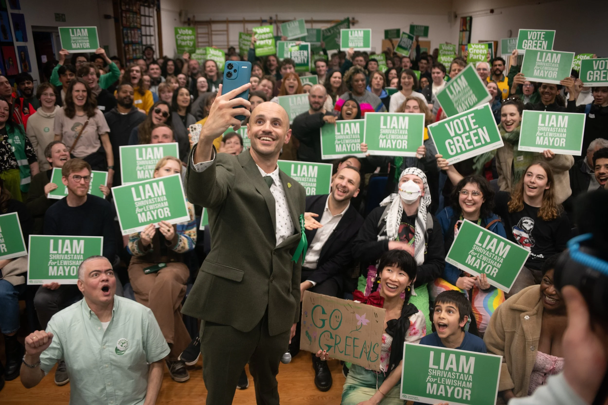 Man taking a selfie with a large crowd holding green campaign signs supporting Liam Shrivastava for Lewisham Mayor, in an indoor community event, with diverse attendees smiling and cheering.