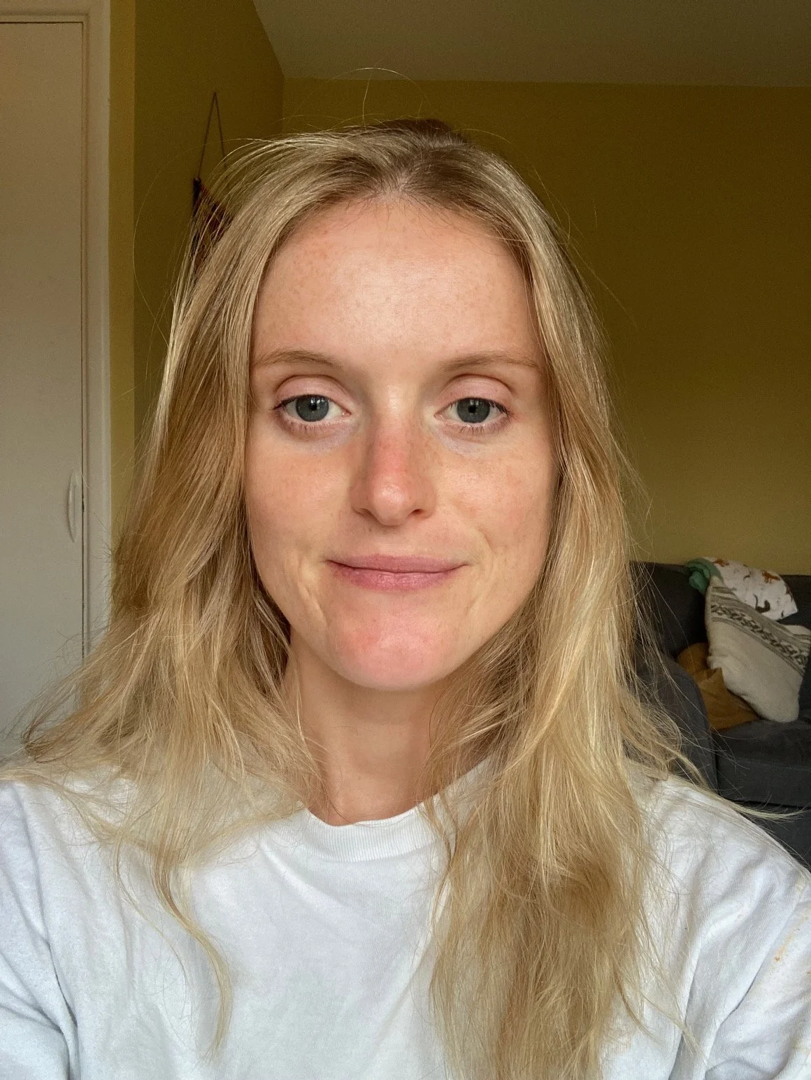 A selfie of a woman with blonde hair, fair skin with freckles, wearing a white shirt, in a room with a yellow wall and a dark couch with pillows.