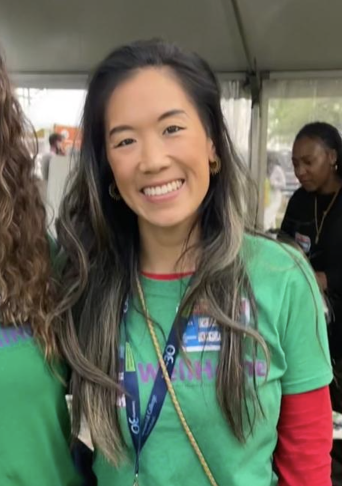 A young Asian woman with long wavy black and gray hair, smiling, wearing a green T-shirt with purple and blue text, a red long-sleeve shirt underneath, gold hoop earrings, and a lanyard around her neck, standing inside a tent.
