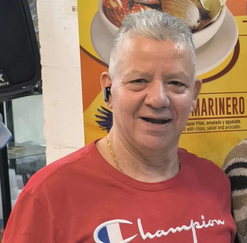A man with short gray hair, wearing a red Champion t-shirt and a gold chain, smiling at the camera. He has an earpiece in his right ear. There is a yellow and orange food poster in the background.