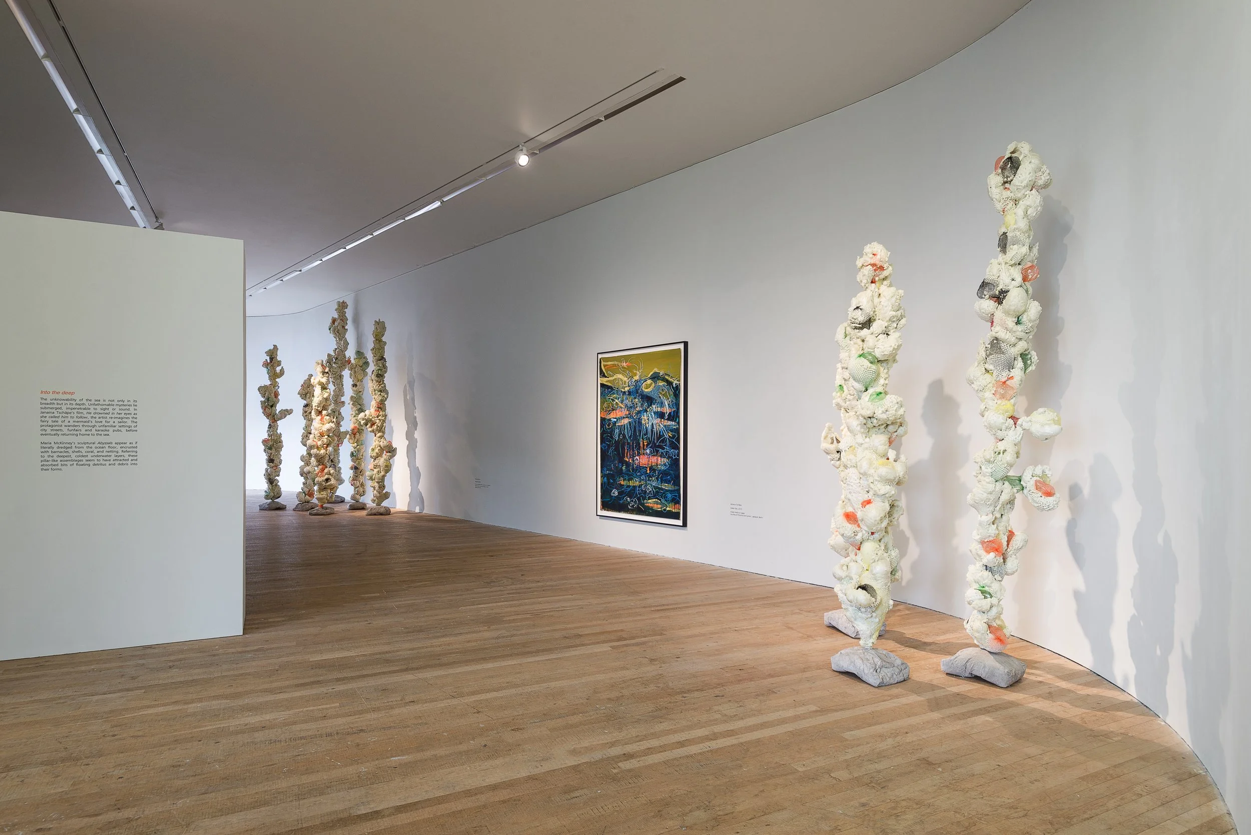 An art gallery with wooden floors and white walls displaying sculptural installations and framed artwork. There are tall abstract sculptures made of mixed materials and a colorful framed painting on the wall.