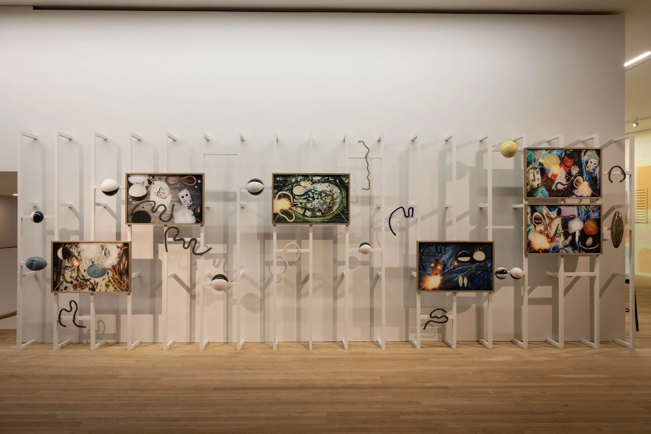 Contemporary art installation with a white wall featuring framed digital screens displaying abstract, colorful art, mounted on white structured shelves with floating spheres and bent wire sculptures casting shadows on the wall.