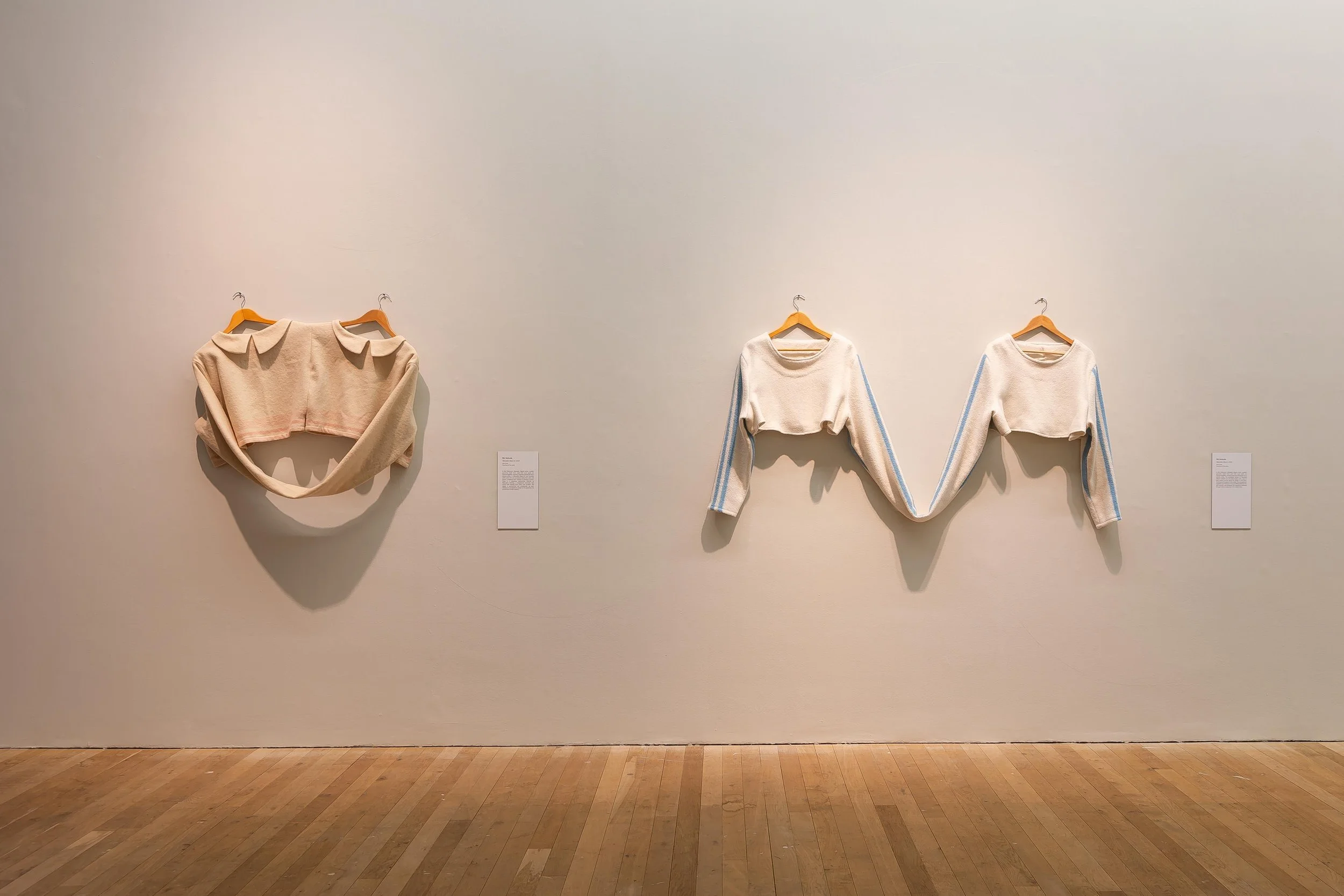 Art installation featuring three clothing pieces on a white gallery wall: a beige cropped sweater with collar on the left, a cream-colored sweater with blue stripes on the sleeves in the middle, and a similar cream sweater with blue stripes on the sl