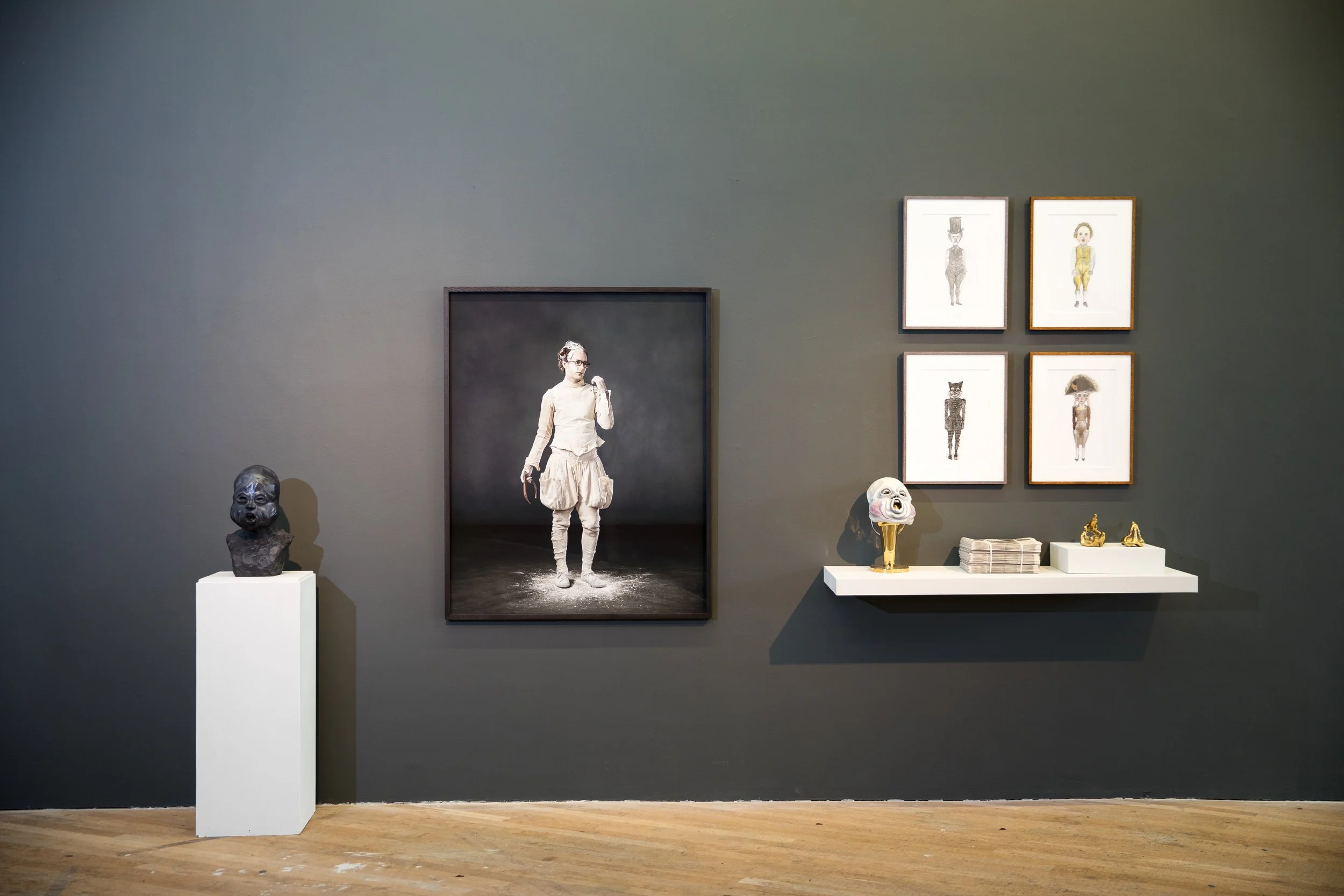 An art gallery wall displaying sculptures and framed sketches, including a bust of a child's head on a white pedestal, a portrait photograph of a woman in vintage clothing, and a colorful animal mask on a stand, with framed sketches of humanoid figur