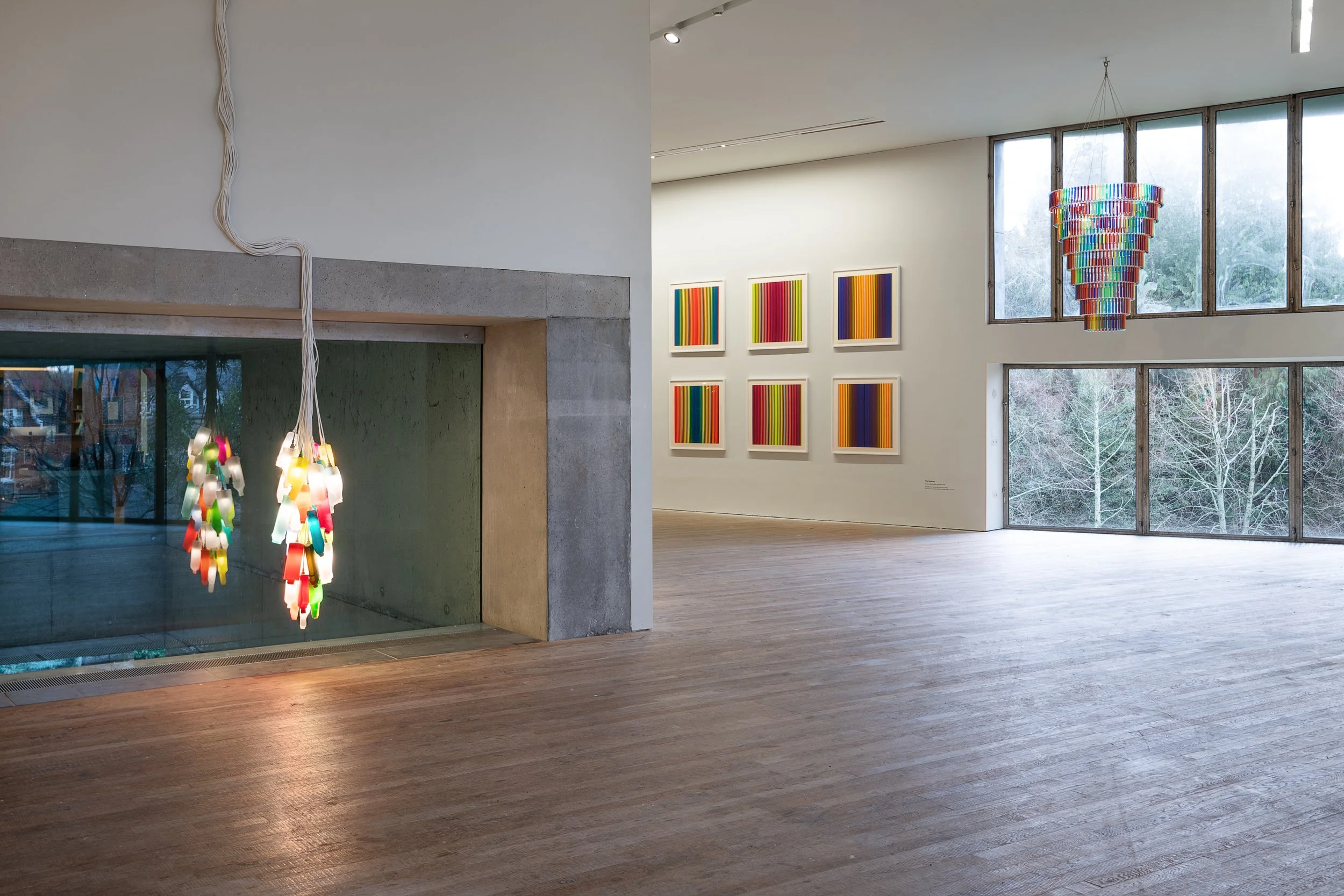 A modern art gallery with colorful abstract paintings on the wall, unique hanging light fixtures, and a large window showing trees outside.