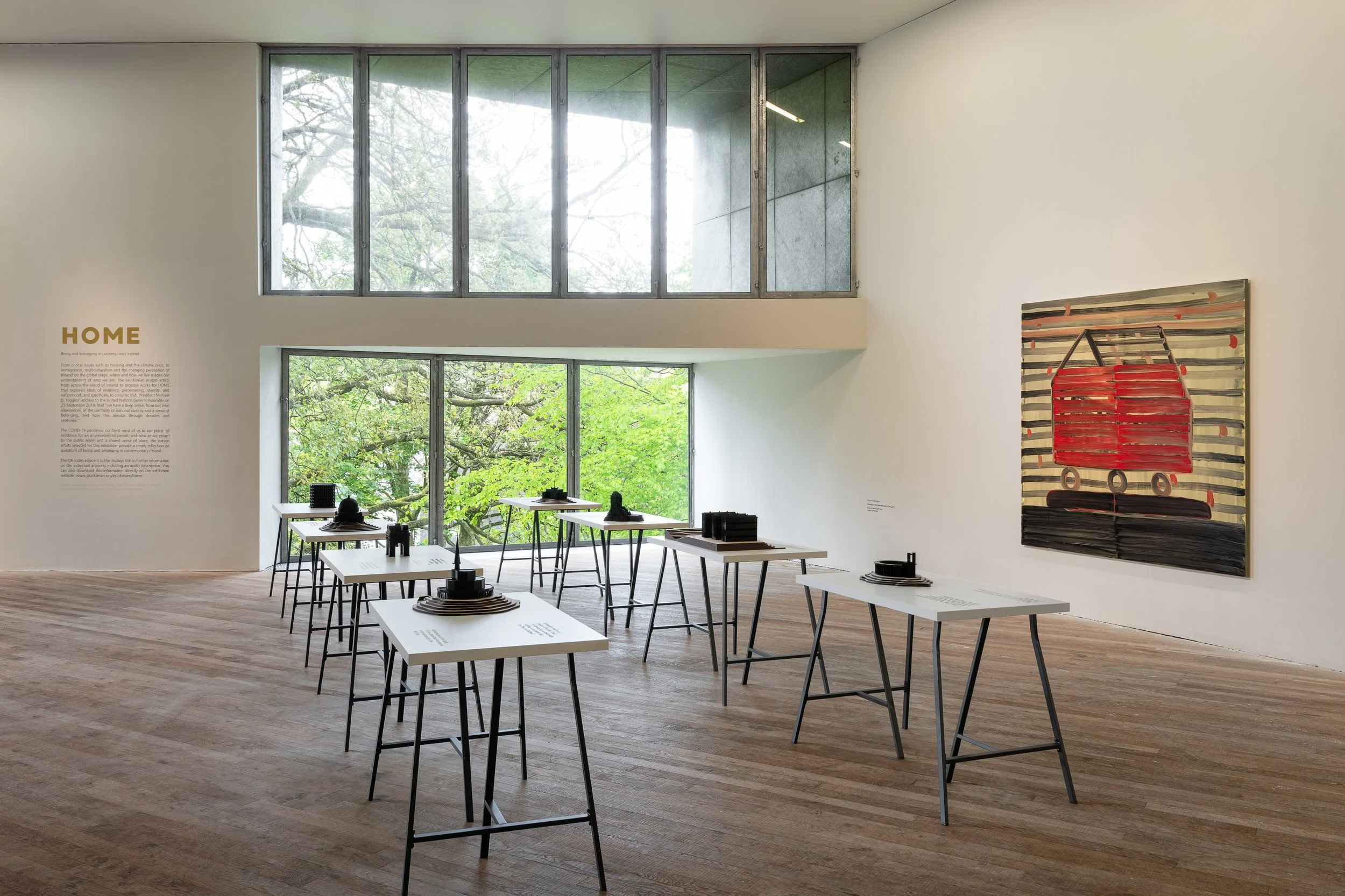 An art gallery with white walls and wooden flooring, featuring a large window with green trees outside. There are several white tables with black objects on top, arranged in the middle of the room. On the right wall, there is an abstract painting of 