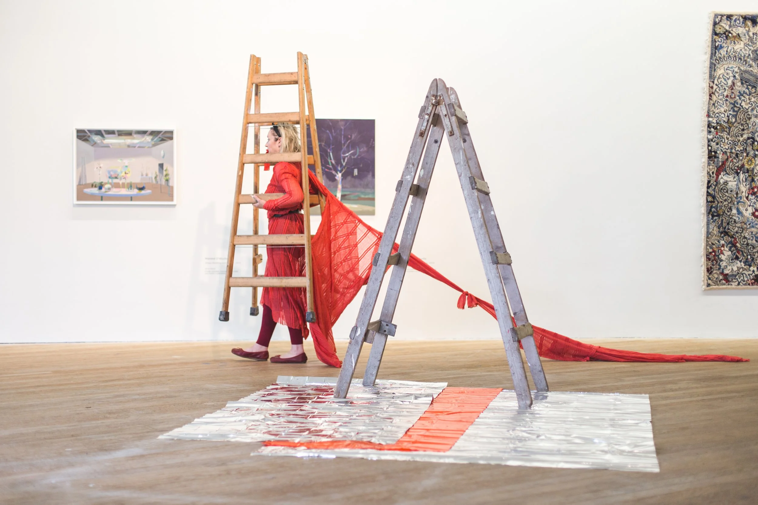 An art installation featuring a woman in a red dress with a red netted fabric attached, standing on a wooden ladder. The setup includes a metallic tarp on the floor with a red and orange pattern, a second ladder, and artworks hanging on the gallery's