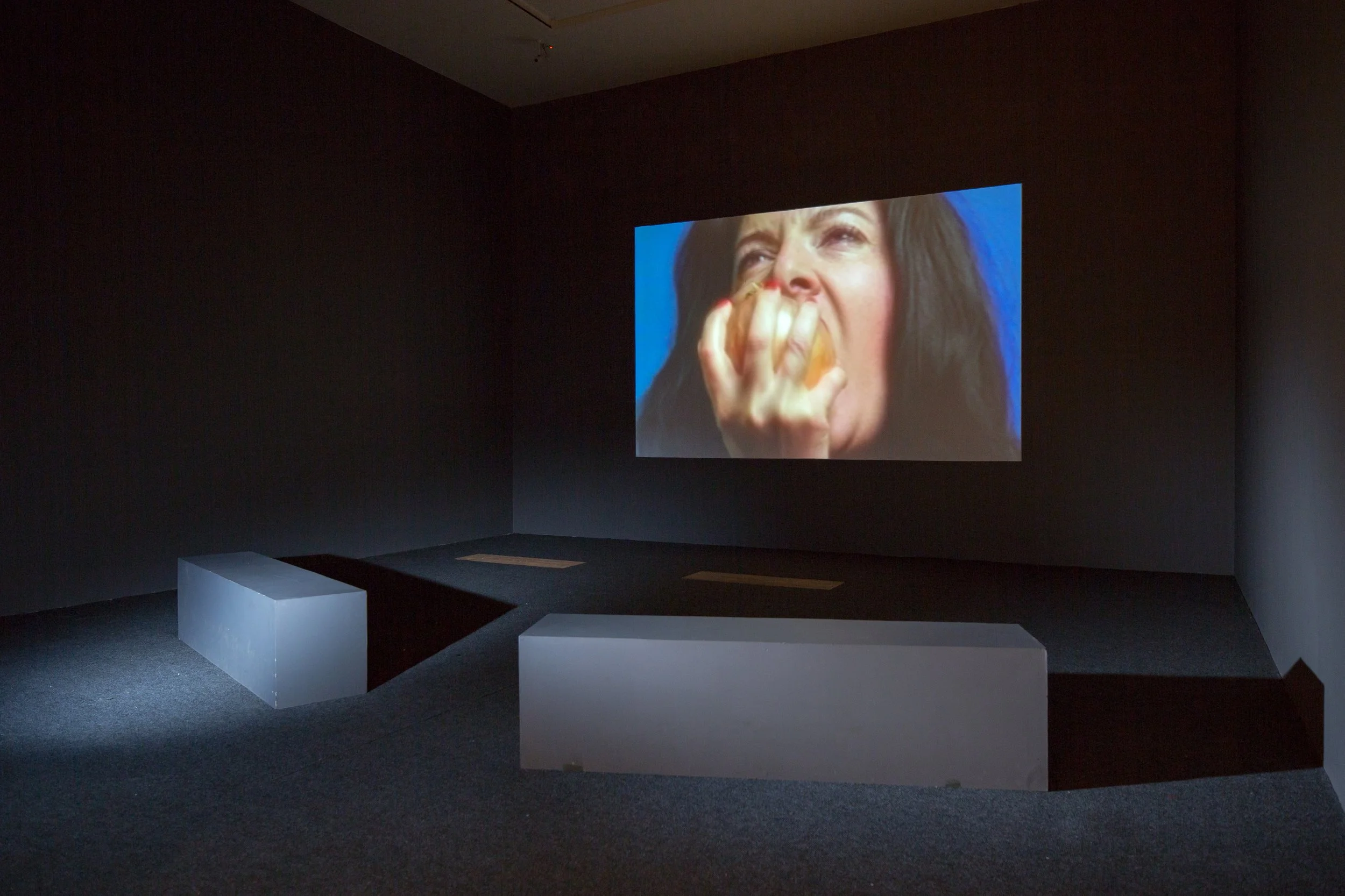 A dark gallery room with a large screen displaying a close-up of a woman with long dark hair; she has a pensive expression and is holding her hand to her face.