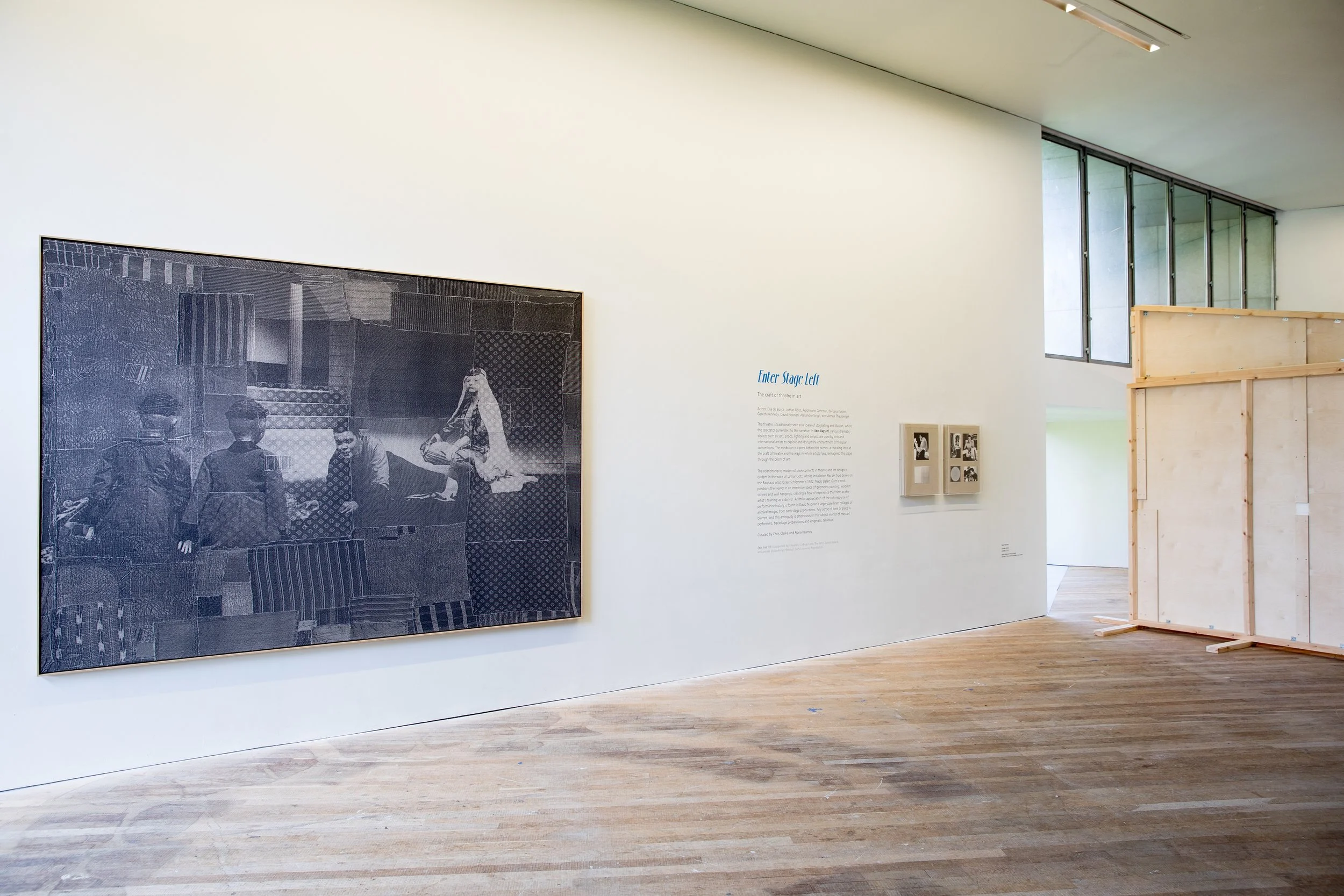 An art gallery with white walls and wooden flooring, featuring a large black and white photograph of three men and a woman in traditional attire on the left side, and a text panel titled "Enter Stage Left" on the right side. There are also two small 