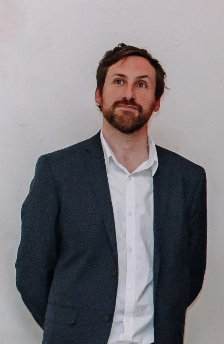 A man in a dark suit jacket and white shirt standing against a plain white wall.