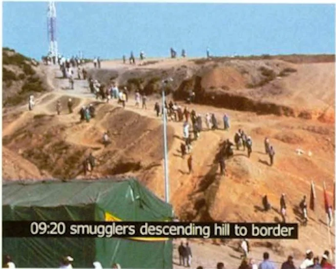 People descending a hill toward a border in a desert landscape, with tents and facilities nearby.