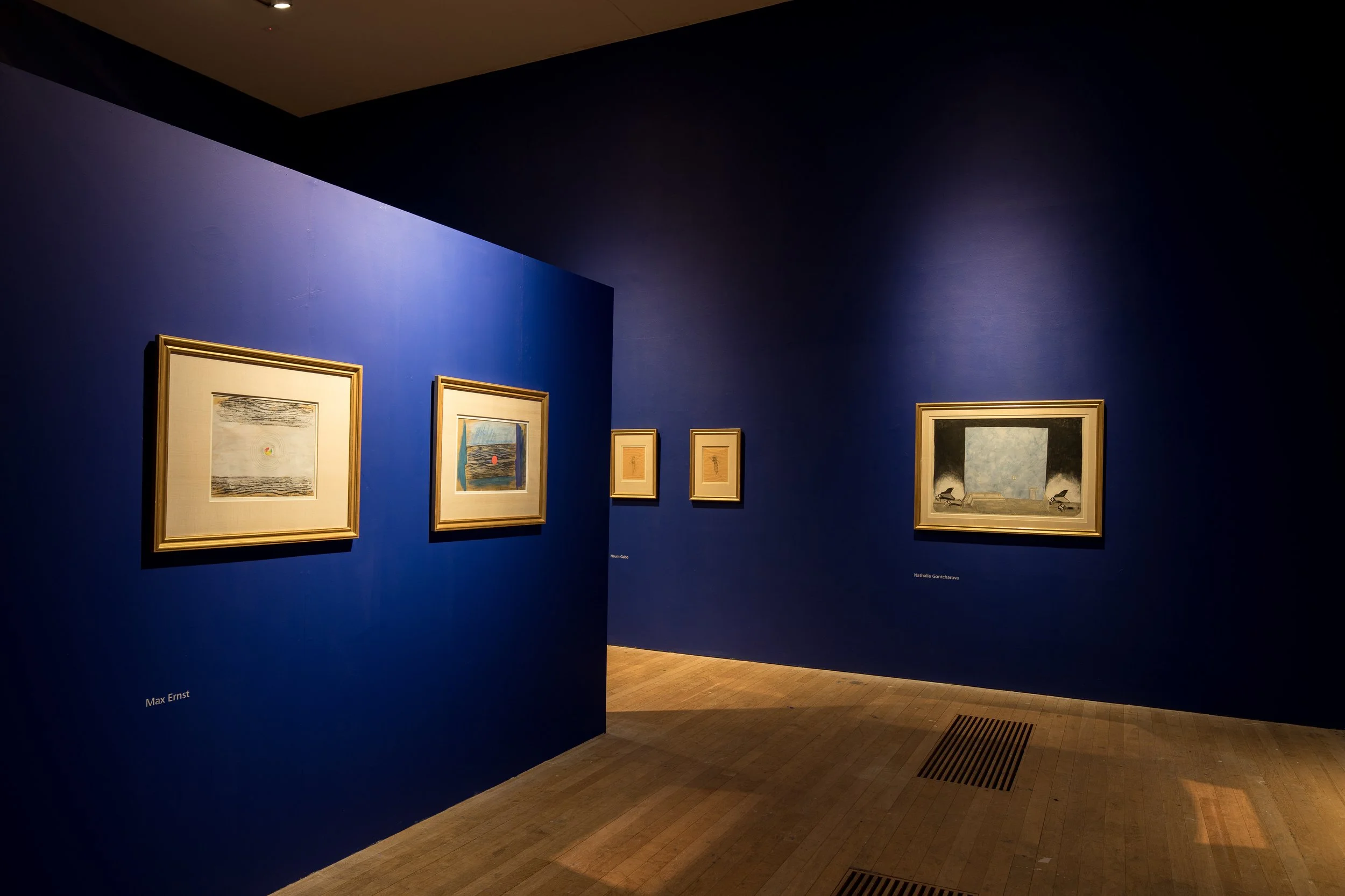 An art gallery with blue walls displaying framed paintings and drawings, including artworks by Max Ernst, Naum Gabo, and Natalia Goncharova, illuminated with spotlights on a wooden floor.