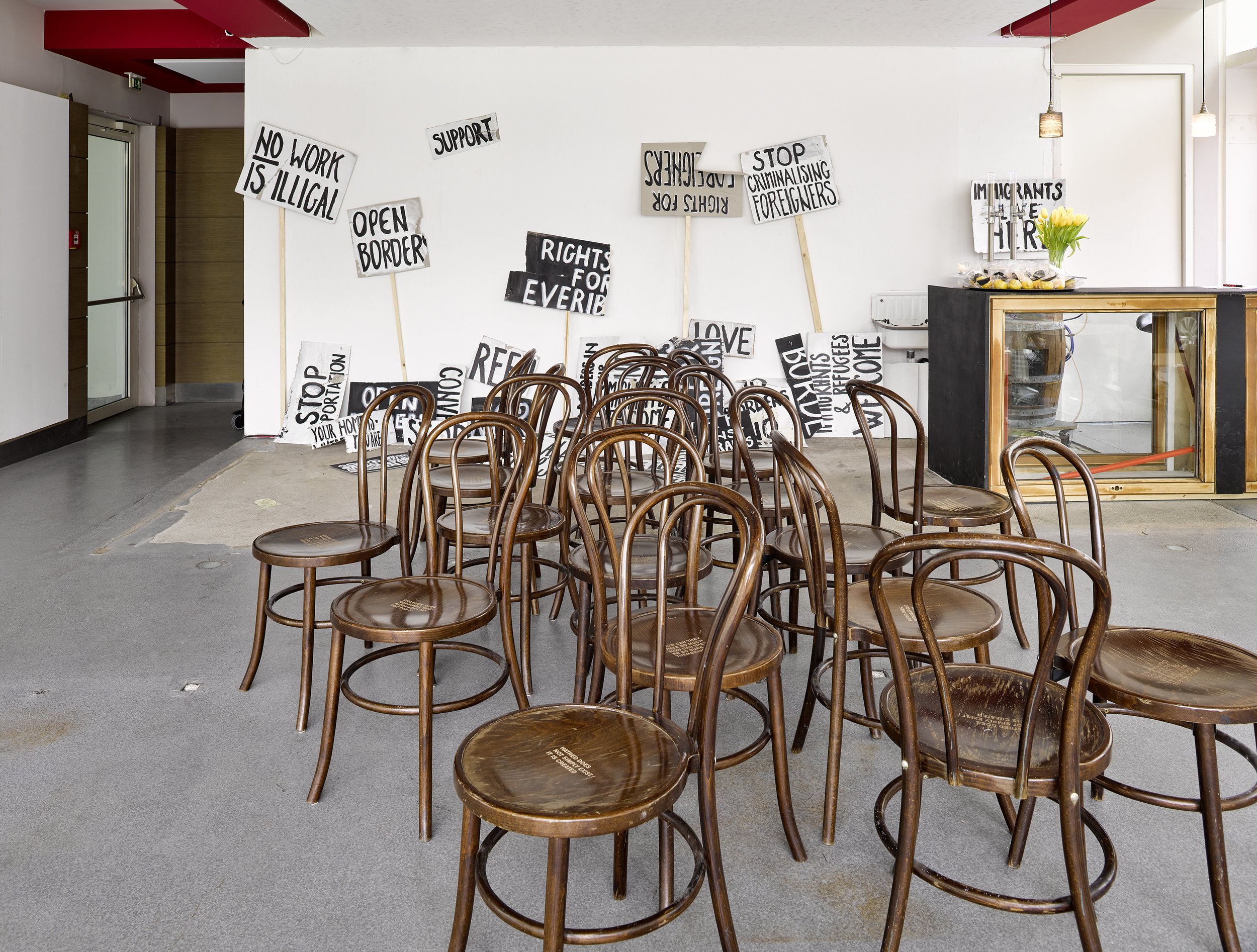 Empty room with scattered wooden chairs and protest signs on the wall, including messages like "No work is illegal," "Stop criminalizing foreigners," and "Support".
