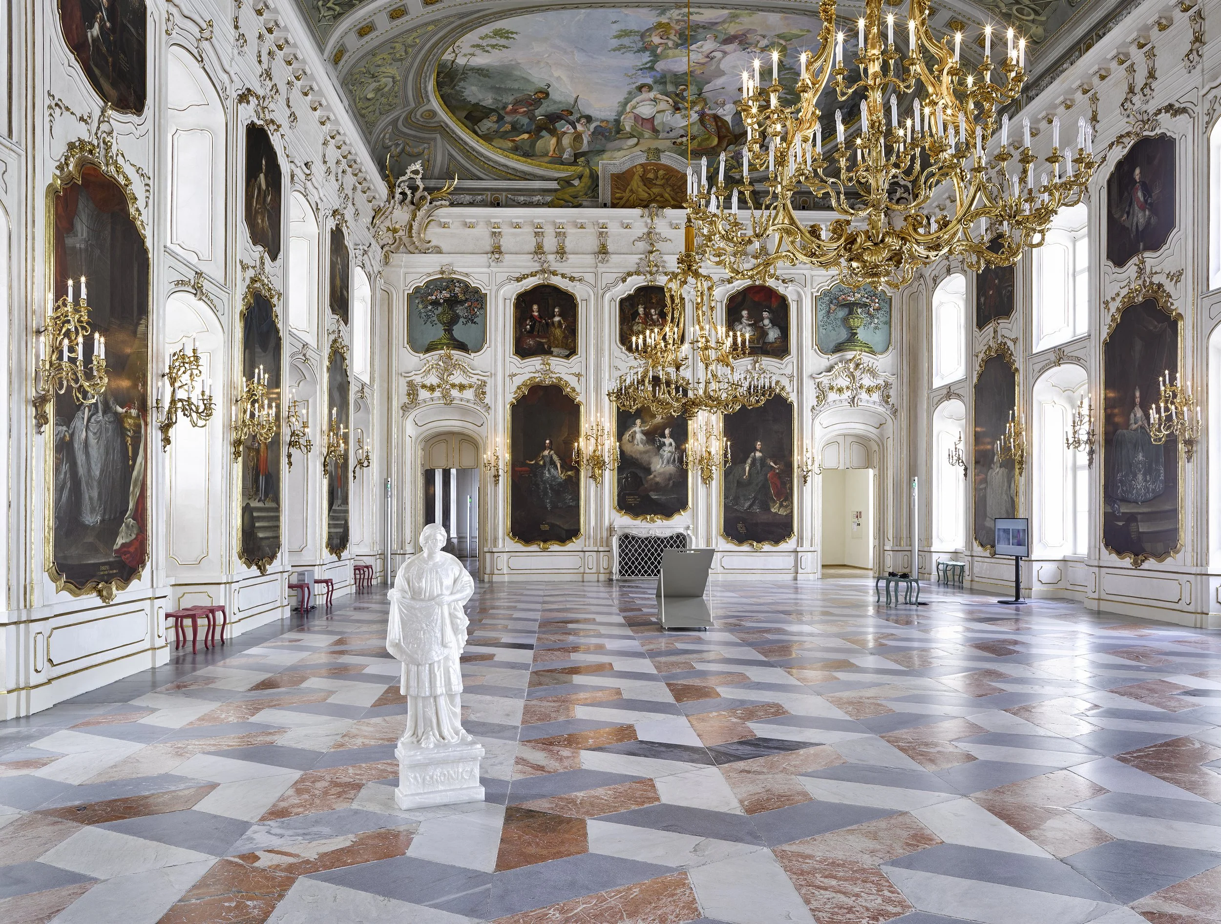 A grand hall with high arched windows, ornate gold chandeliers, large paintings with gold frames on white paneled walls, a marble floor with geometric pattern, a white statue, and digital display screens.
