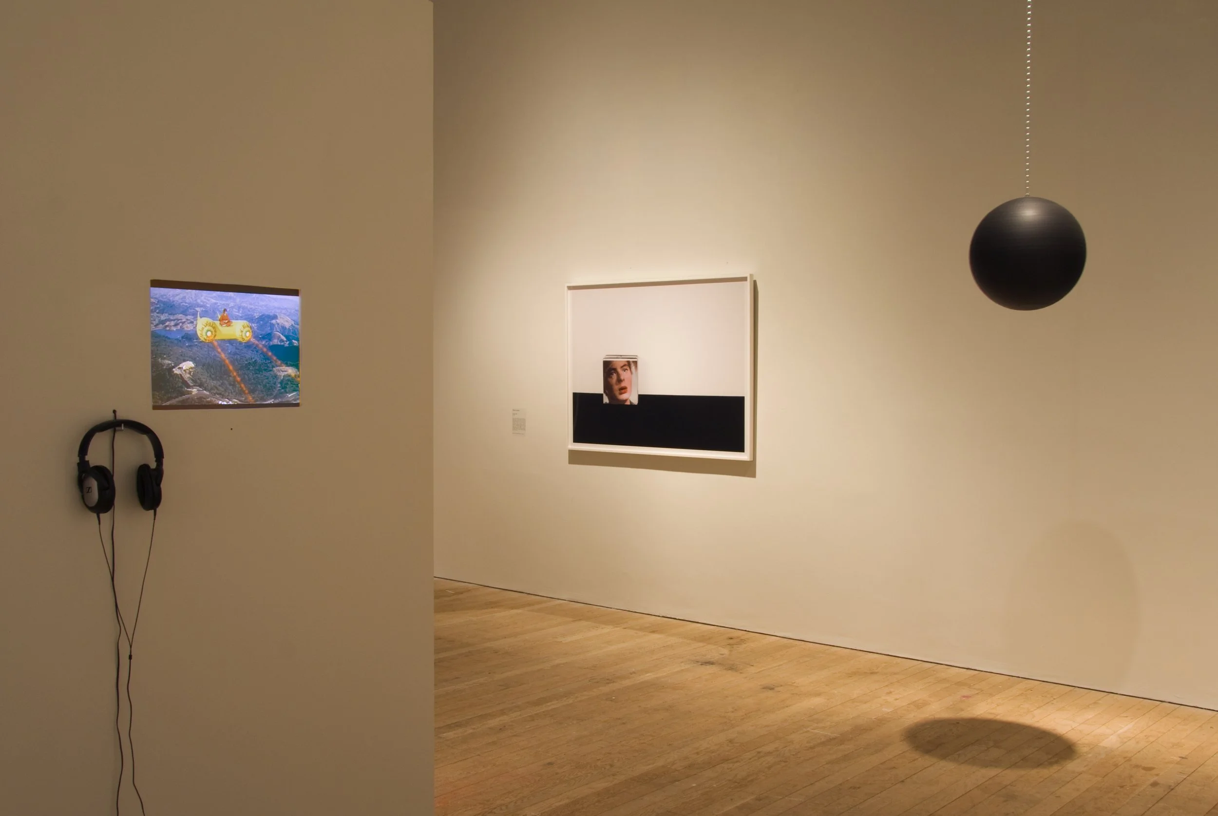 An art gallery display with a framed artwork featuring a woman's face and black and white backgrounds, a small screen showing a yellow vehicle in a landscape, black headphones hanging on the wall, and a spherical black object hanging from the ceiling