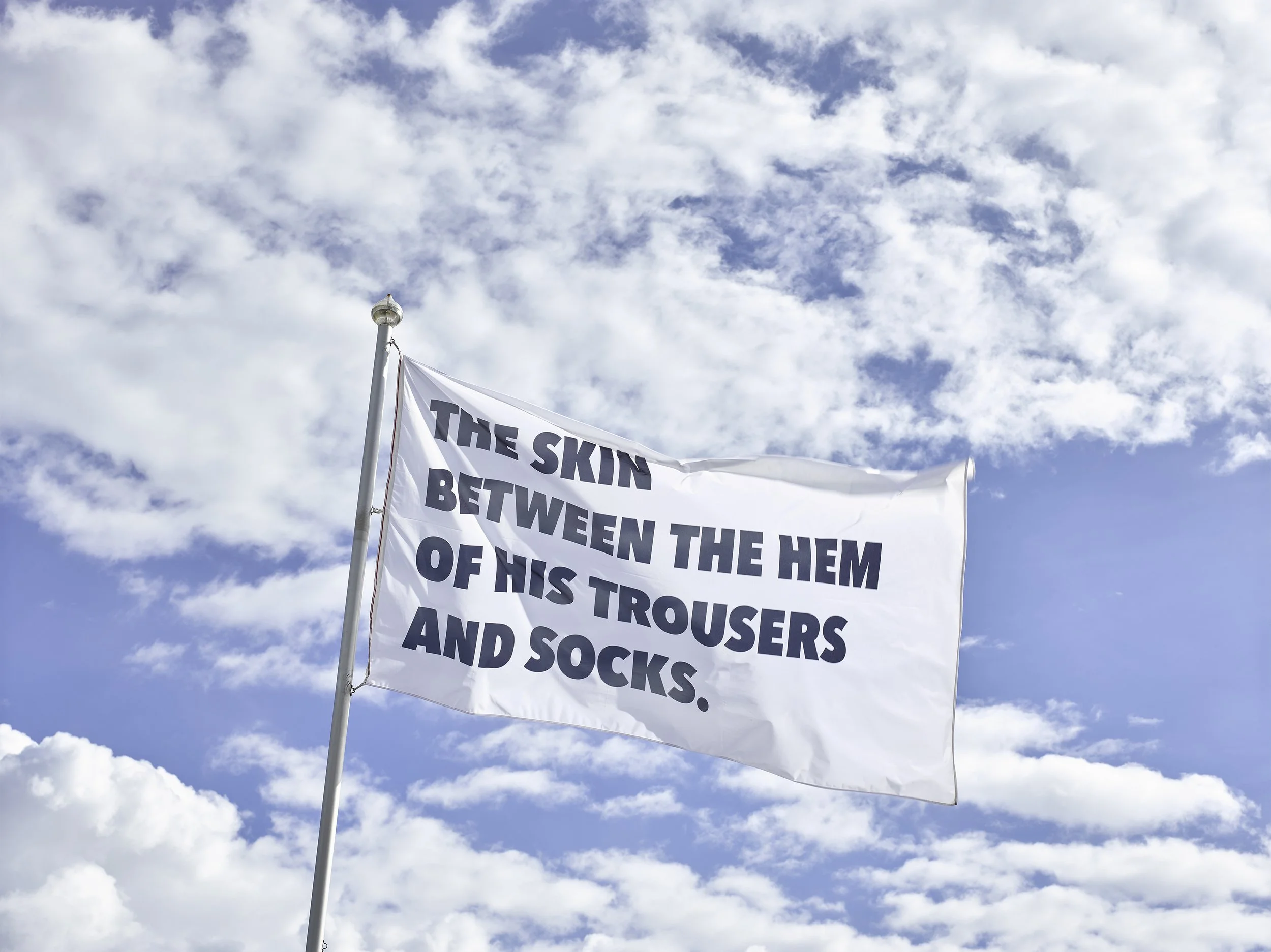A white flag with black text flying against a cloudy blue sky. The text reads: 'The skin between the hem of his trousers and socks.'