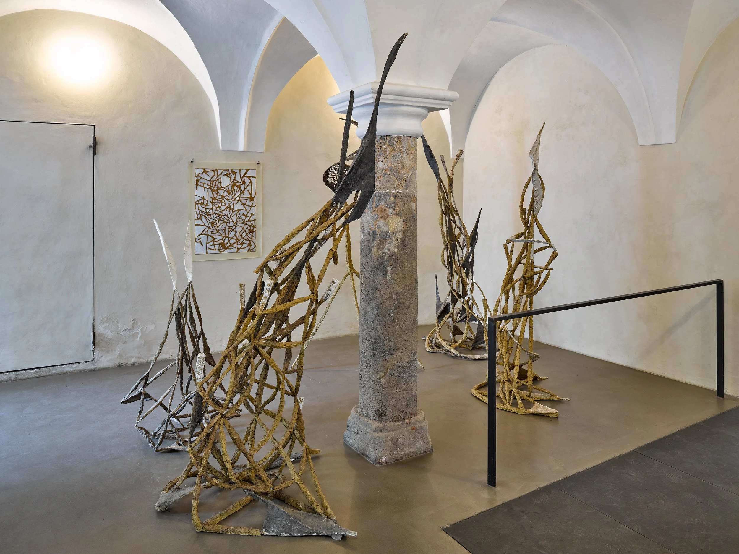 Art installation featuring abstract sculptures of elongated, intertwined forms made of textured materials, displayed around a central stone column in a white-walled gallery with arched ceilings.