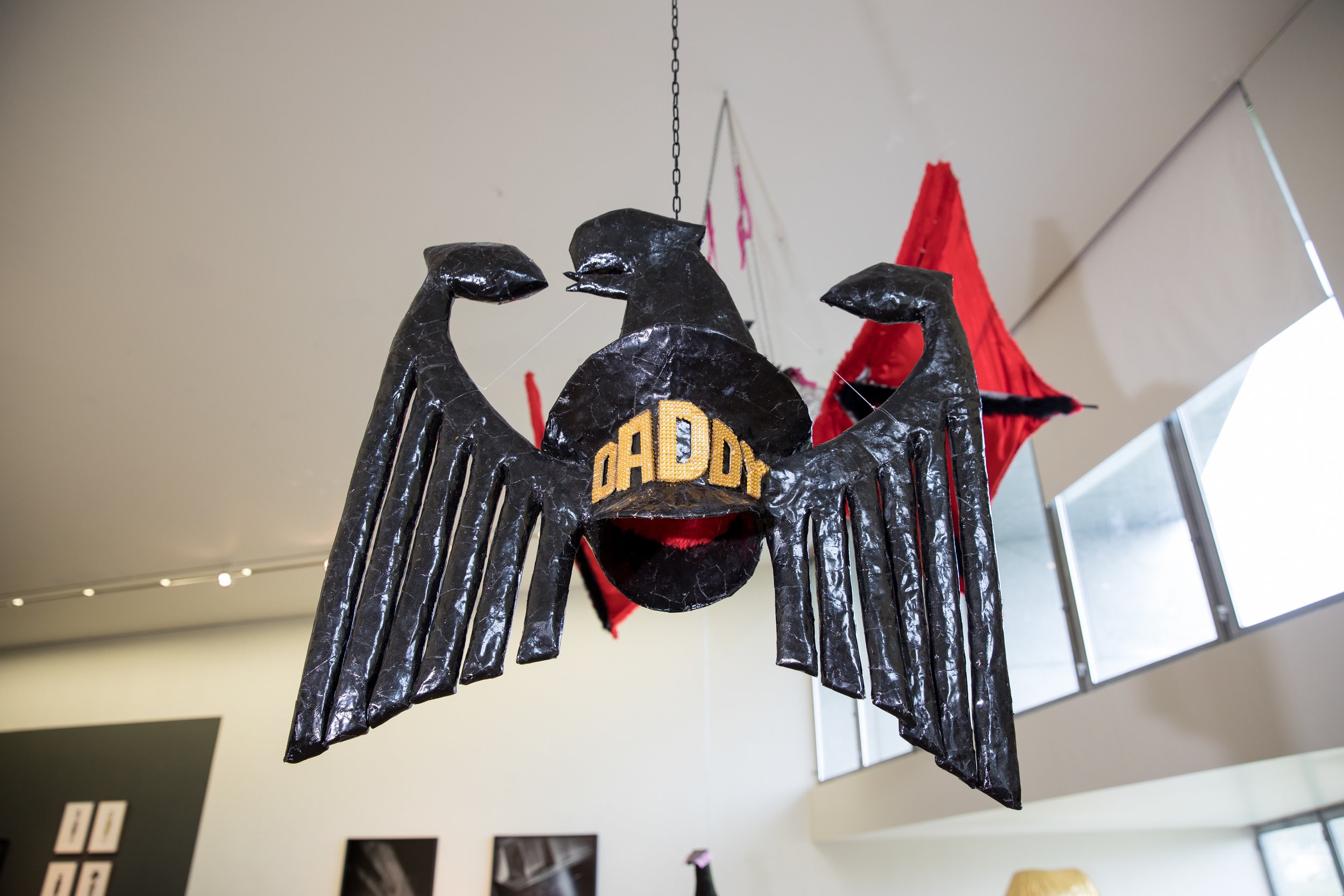 A black hanging sculpture of an eagle with outstretched wings, featuring the word 'DADDY' on its chest, in an indoor art gallery with windows and other artwork visible in the background.