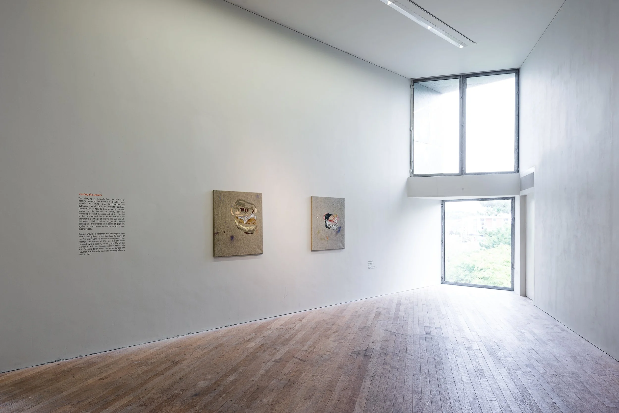 An art gallery with white walls and wooden flooring, featuring two paintings and a wall text panel. A large window with a geometric frame lets in natural light.