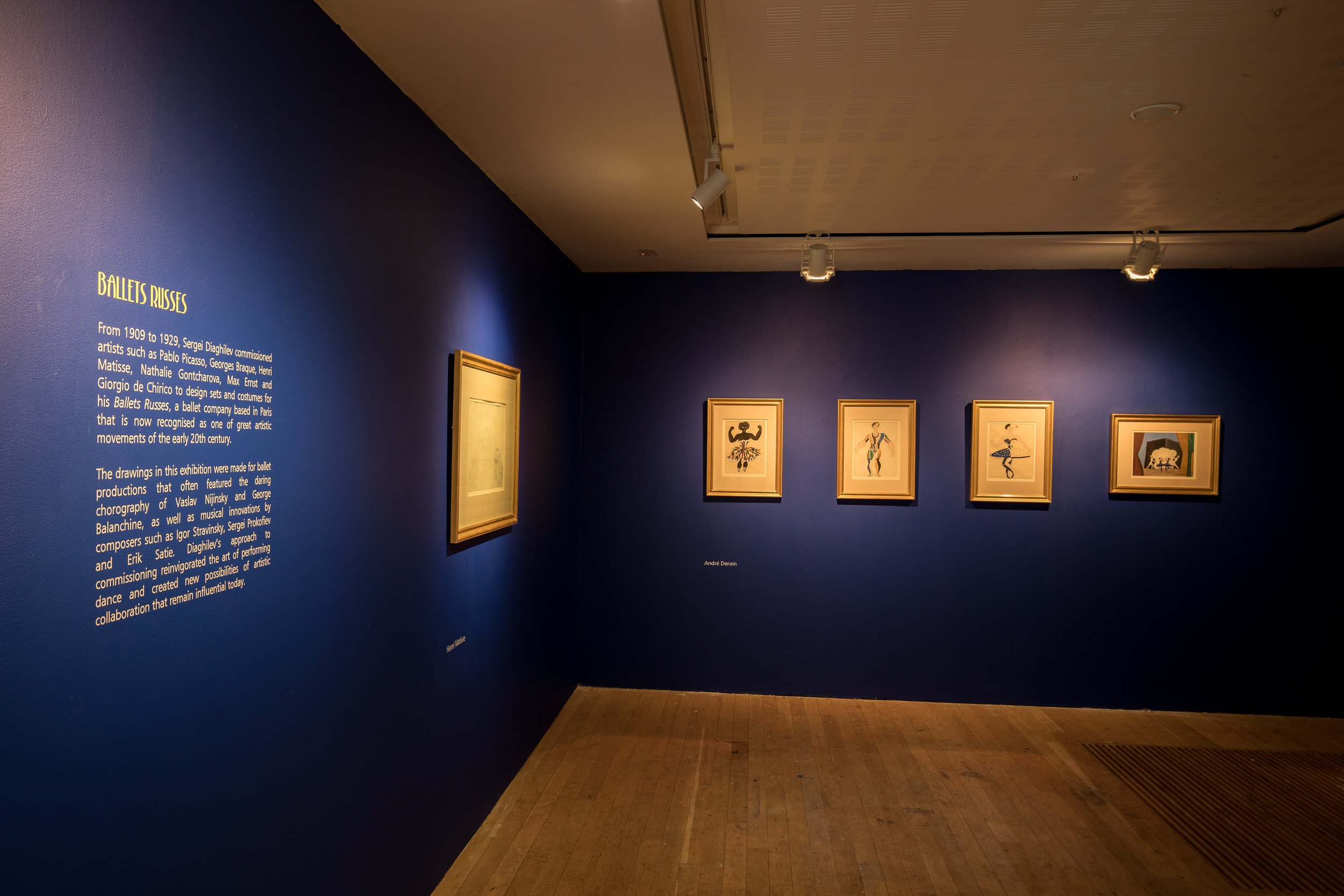 An art gallery with blue walls displaying framed artwork and informational text panel about Ballets Russes.