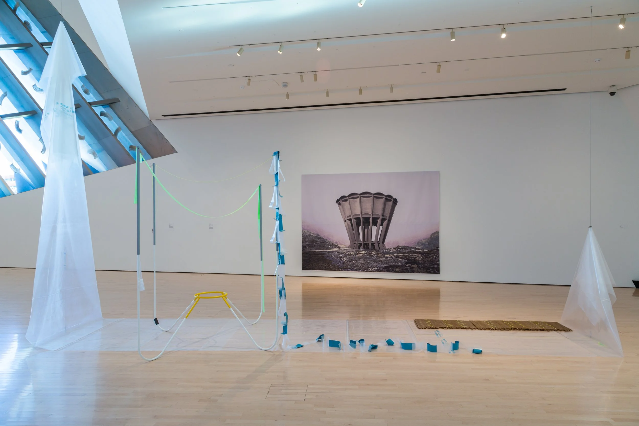 Contemporary art installation in a gallery with a large abstract sculpture, colorful ribbons, white drapes, and a large landscape photograph of a tower-like structure in the background.