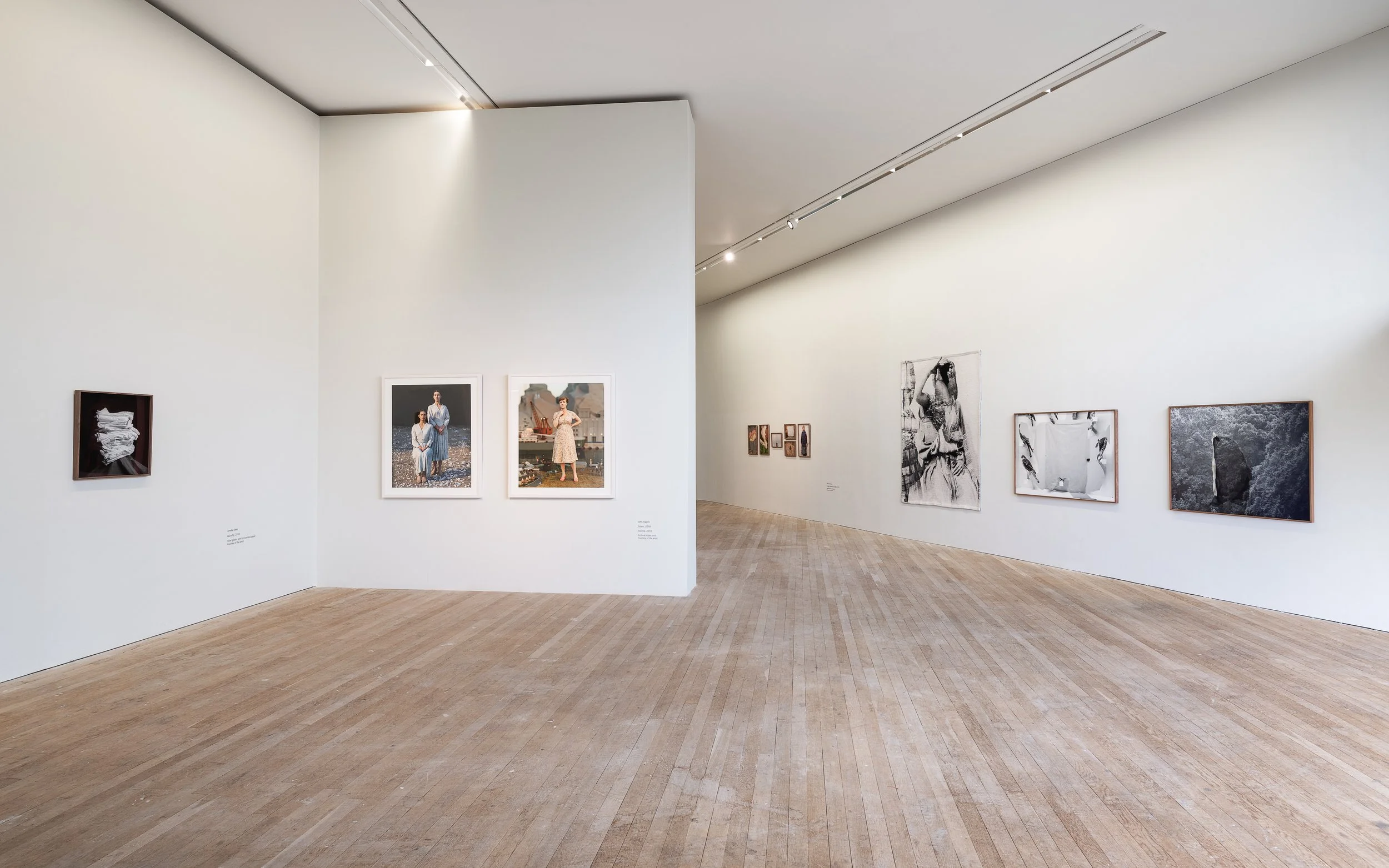 An art gallery with white walls displaying various framed photographs and art pieces. The gallery has wooden flooring and high ceilings with track lighting.