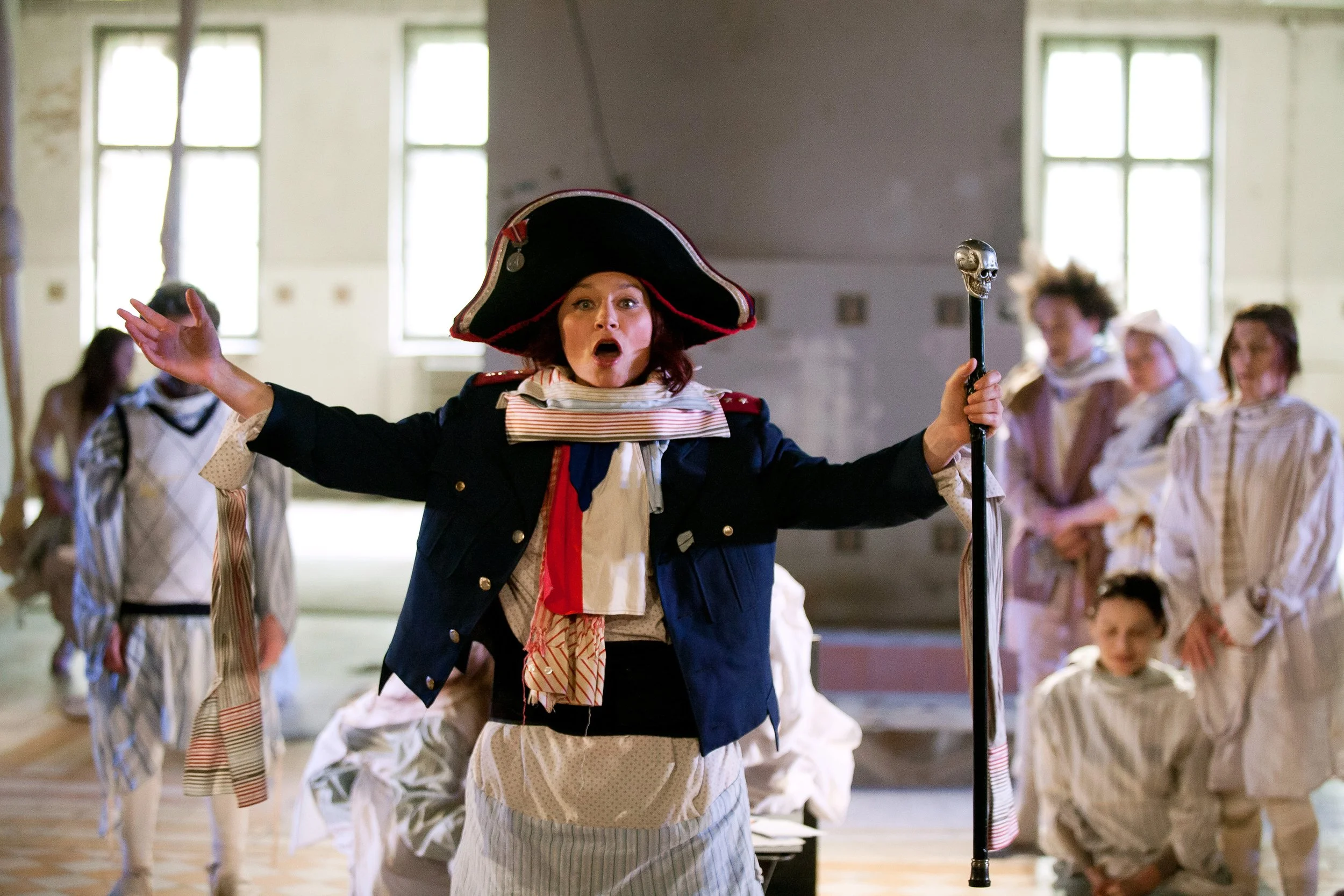 A woman dressed as Napoleon Bonaparte performing in a theatrical production with children dressed in period costumes in the background.
