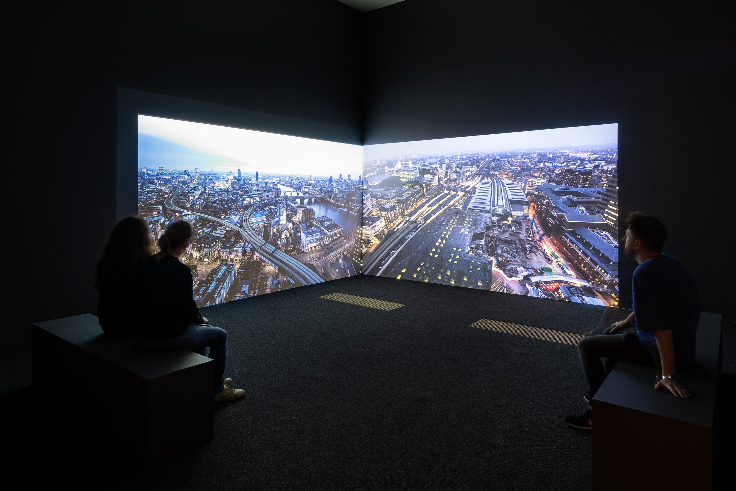 Three people sitting in front of a large panoramic urban cityscape display in a dark room.