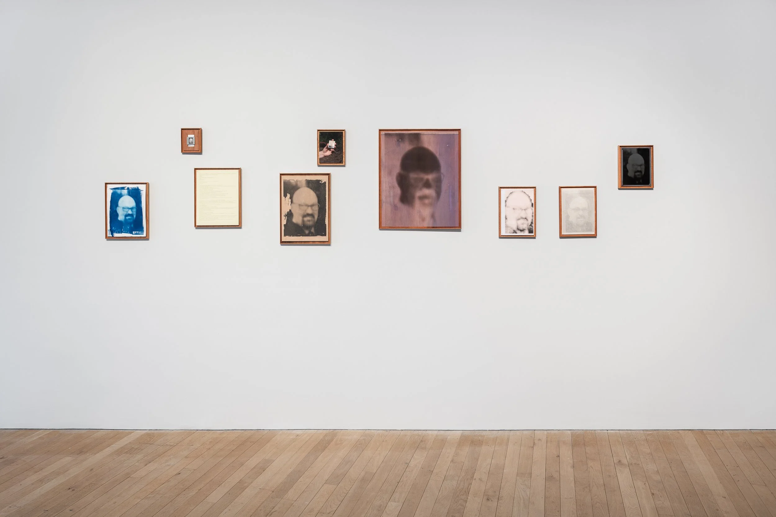 Gallery wall with multiple framed photos and artworks, including a central large portrait of a person's face, on a white wall in an art museum or gallery.