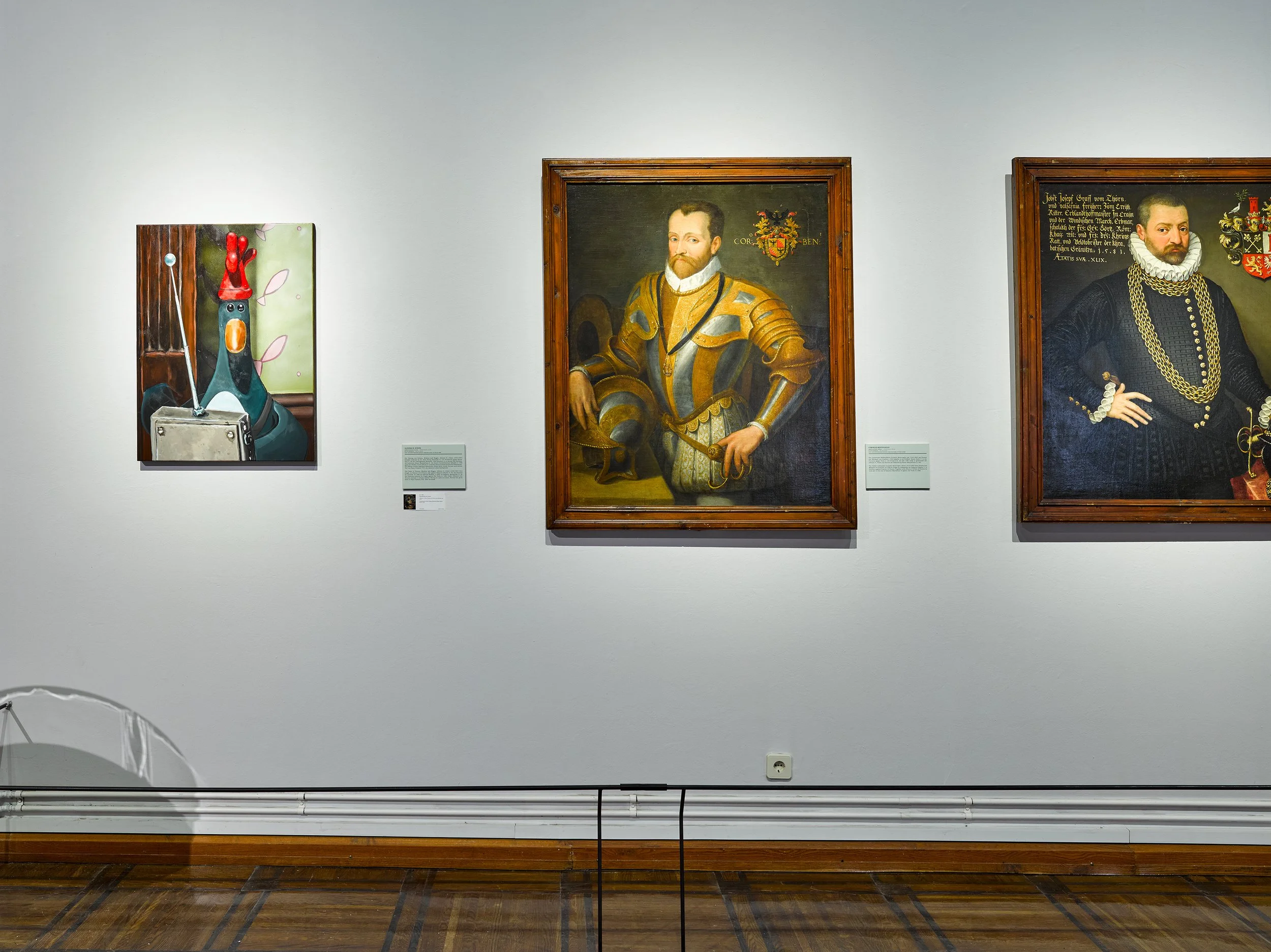 Art gallery wall with three portraits of historical figures and a cartoon chicken with a red party hat, sitting at a desk with a laptop.
