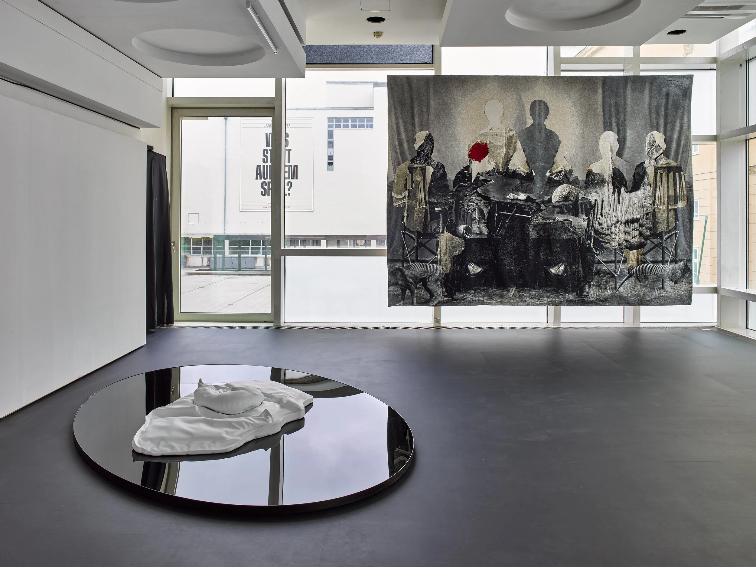 Art gallery space with large black and white hanging artwork, a reflective round floor piece with white fabric and forms, and large windows showing city buildings outside.