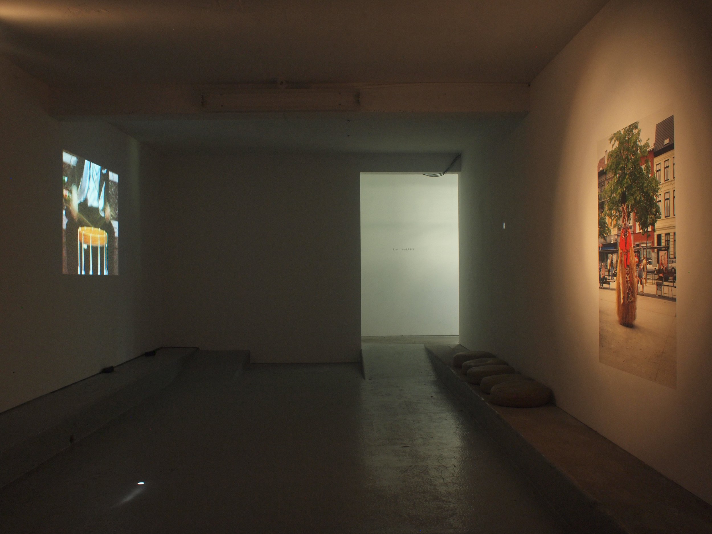 An art gallery with two photographs on the walls. The photo on the left is a close-up of two beer glasses. The photo on the right depicts a city street scene with a person in traditional clothing, trees, and buildings.