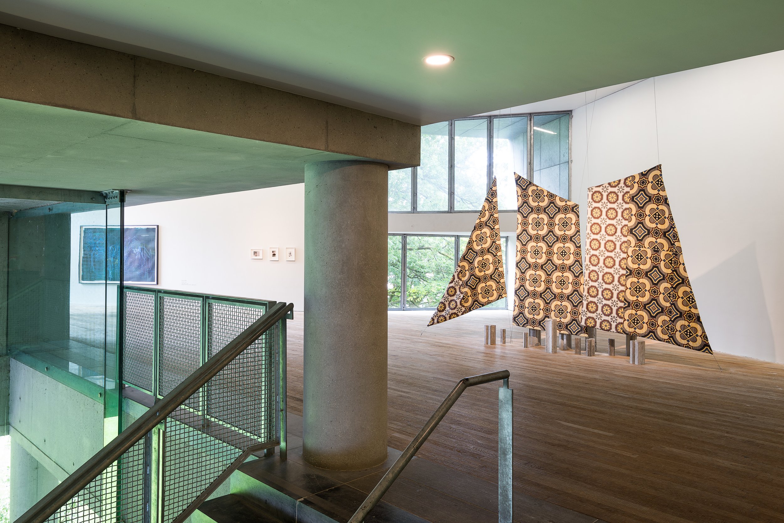 Interior view of an art gallery with patterned fabric sculptures hanging in front of large windows, a wooden floor, and white walls with framed artwork.