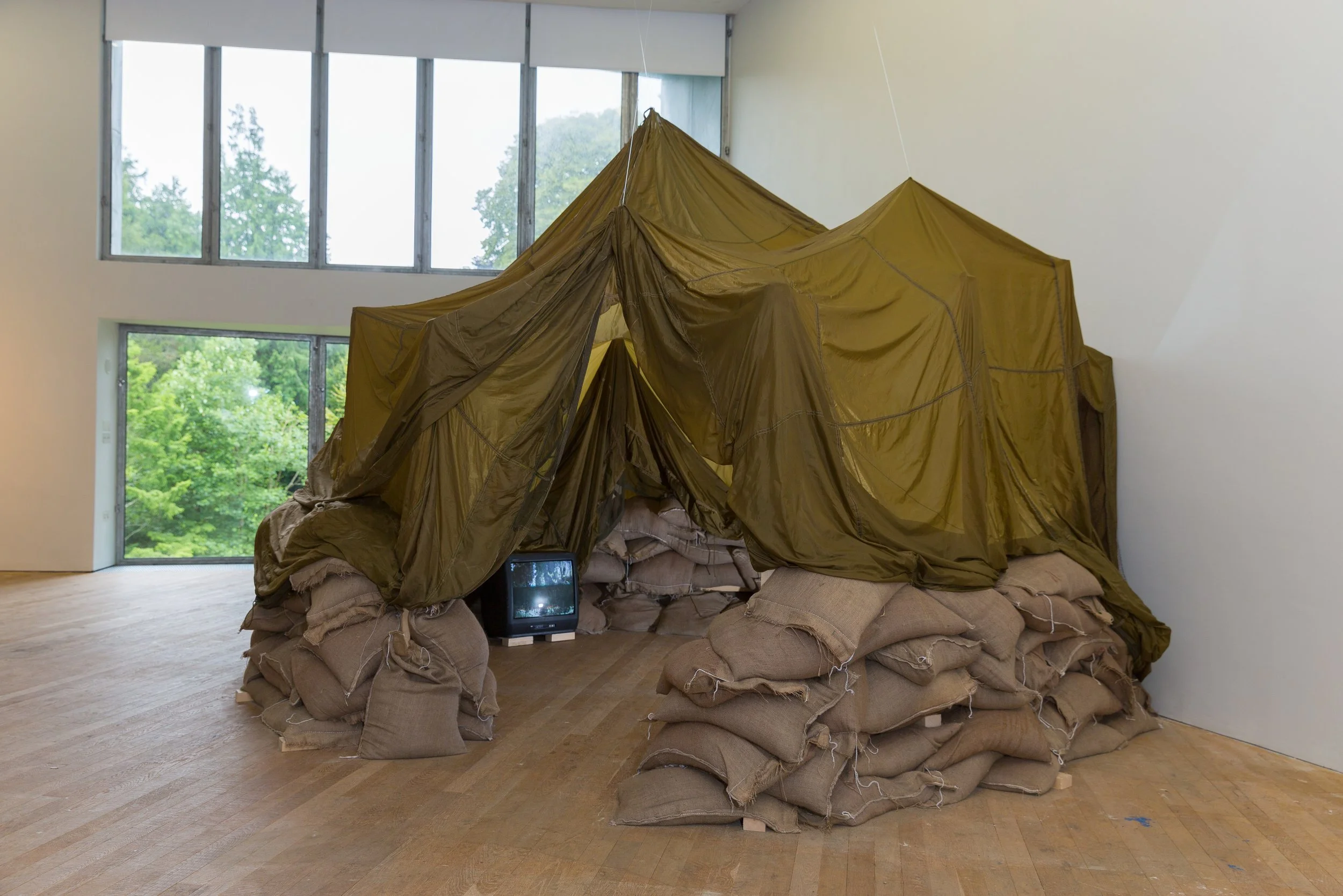 An art installation resembling a makeshift tent made of sandbags, olive drab fabric, and a small vintage television, set inside a modern room with large windows showing green trees outside.