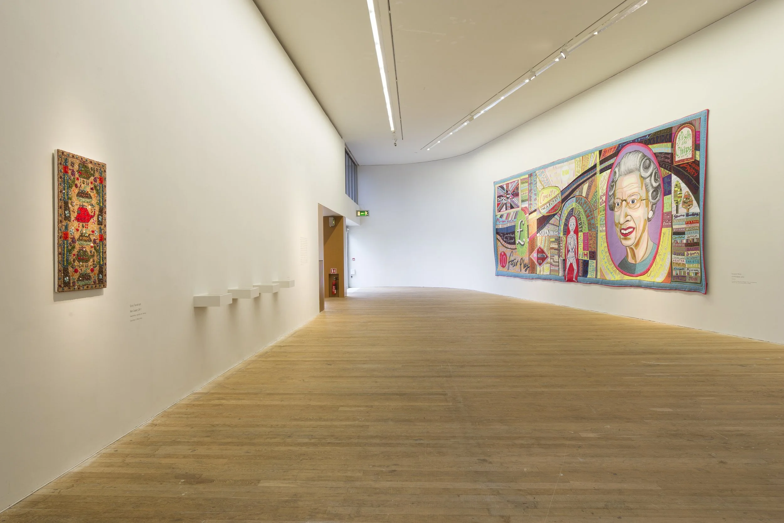 An art gallery with wooden floors and white walls, displaying colorful contemporary paintings, including a portrait of Queen Elizabeth II and a symbolic collage with various graphics.